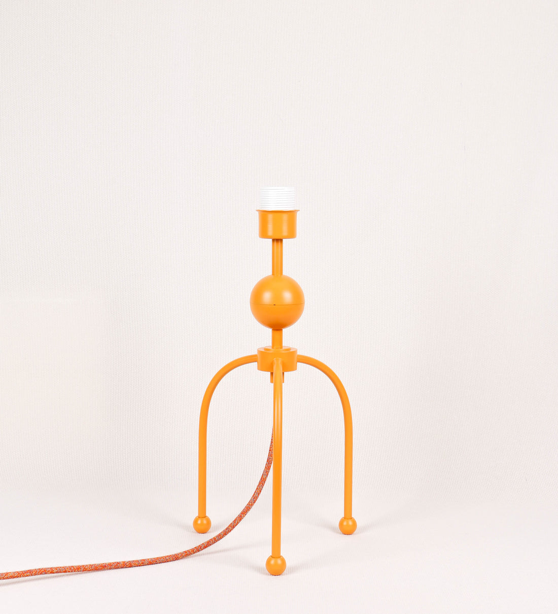 Candeeiro de mesa, de tom laranja-tangerina, com um design moderno e minimalista. O candeeiro tem o corpo com três pés curvados, terminando em pequenas esferas. A estrutura dos pés é interligada por uma peça central esférica. O cabo é revestido têxtil com um padrão de linhas coloridas (laranjas, azuis, amarelas e outras), com interruptor e ficha transparentes.