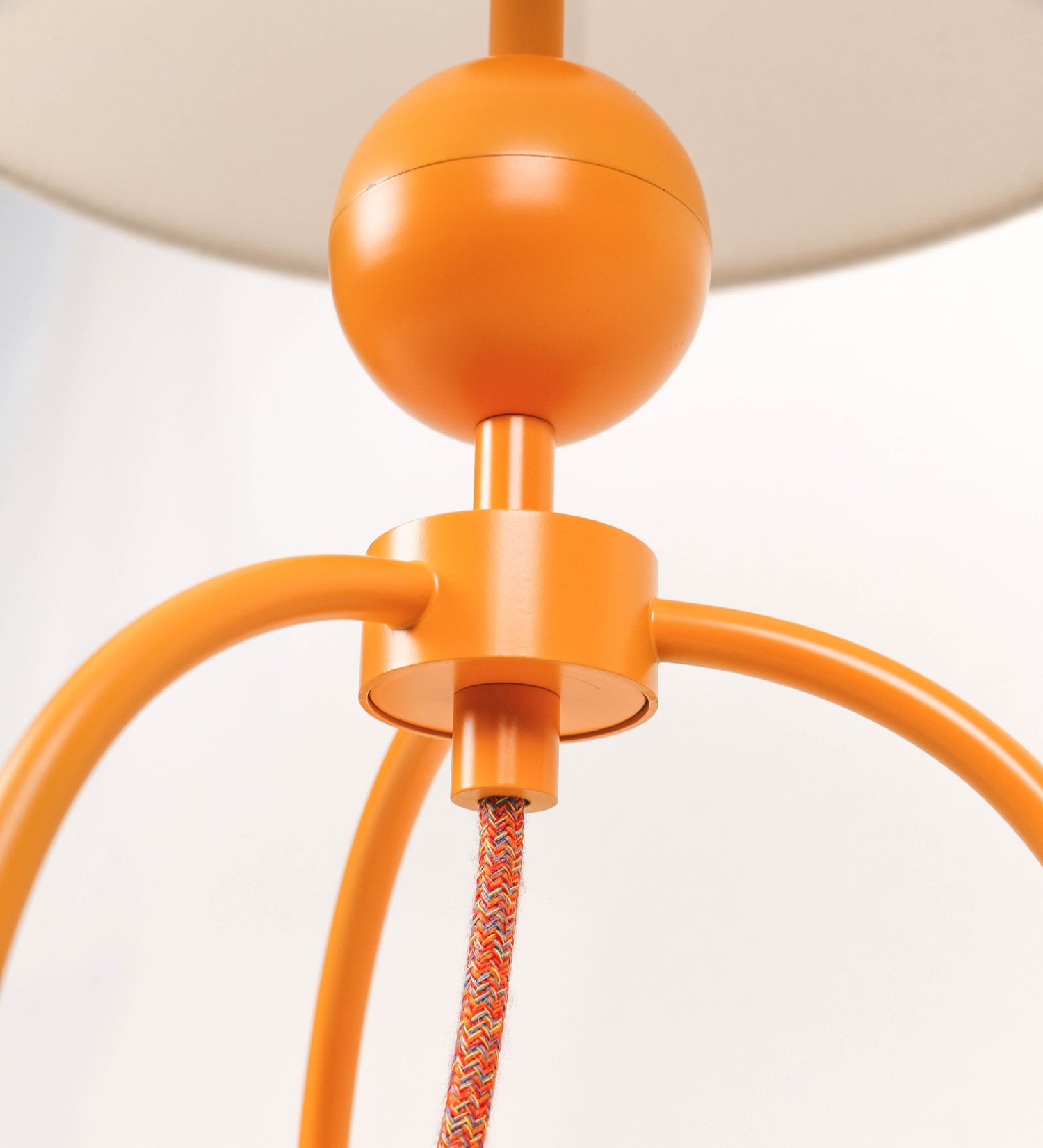 Candeeiro de mesa, de tom laranja-tangerina, com um design moderno e minimalista. O candeeiro tem o corpo com três pés curvados, terminando em pequenas esferas. A estrutura dos pés é interligada por uma peça central esférica. O cabo é revestido têxtil com um padrão de linhas coloridas, com interruptor e ficha transparentes. O abatjour é de forma cónica, em algodão, de tom bege claro com pequenas pintas escuras, semelhante à baunilha.