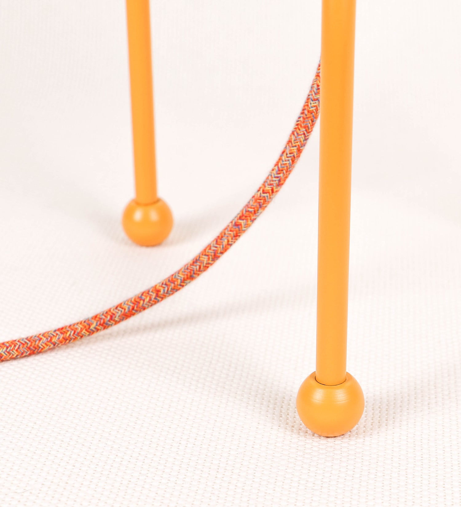 Candeeiro de mesa, de tom laranja-tangerina, com um design moderno e minimalista. O candeeiro tem o corpo com três pés curvados, terminando em pequenas esferas. A estrutura dos pés é interligada por uma peça central esférica. O cabo é revestido têxtil com um padrão de linhas coloridas, com interruptor e ficha transparentes. O abatjour é de forma cónica, em algodão, de tom bege claro com pequenas pintas escuras, semelhante à baunilha.