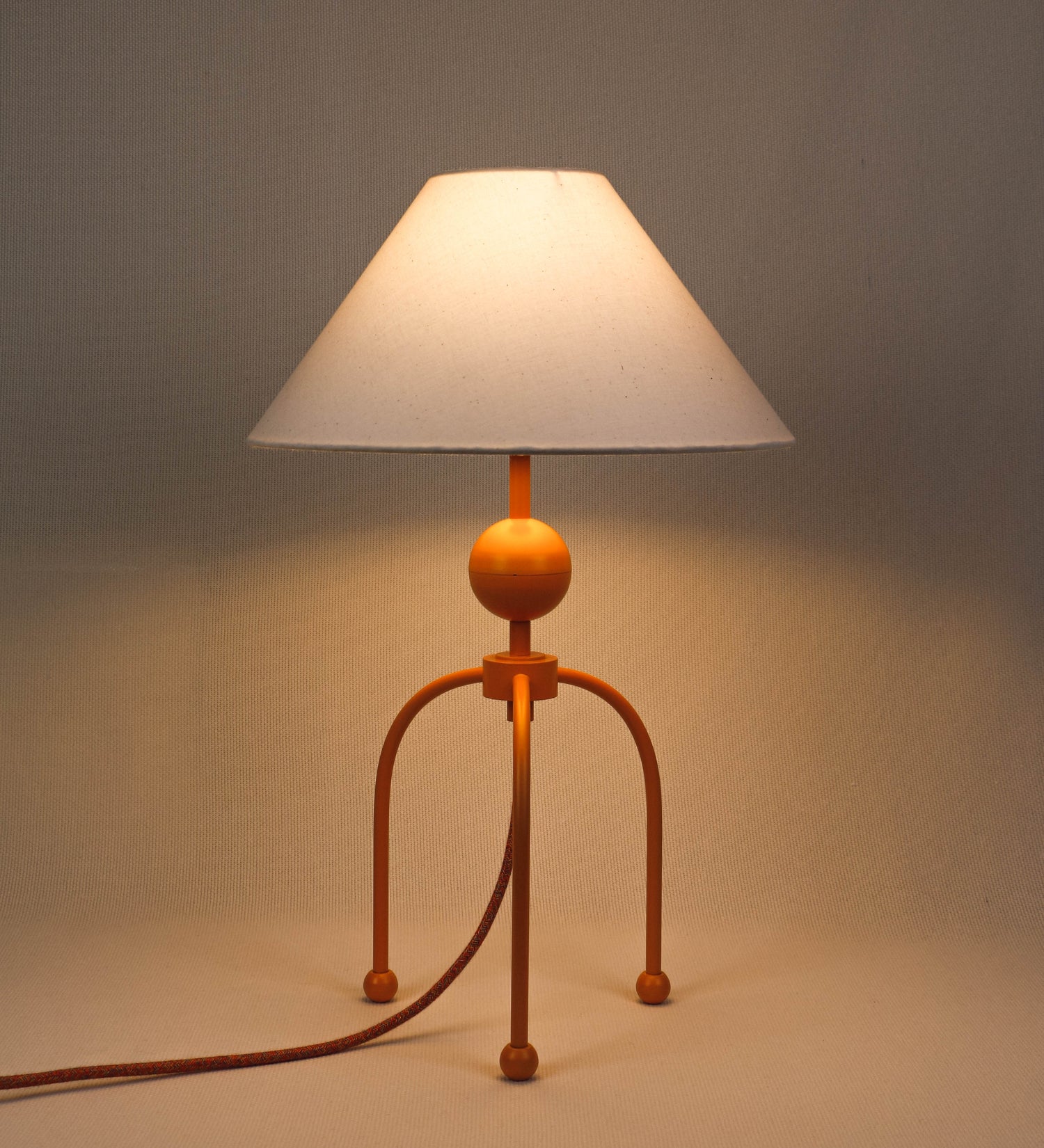 Candeeiro de mesa, de tom laranja-tangerina, com um design moderno e minimalista. O candeeiro tem o corpo com três pés curvados, terminando em pequenas esferas. A estrutura dos pés é interligada por uma peça central esférica. O cabo é revestido têxtil com um padrão de linhas coloridas, com interruptor e ficha transparentes. O abatjour é de forma cónica, em algodão, de tom bege claro com pequenas pintas escuras, semelhante à baunilha.
