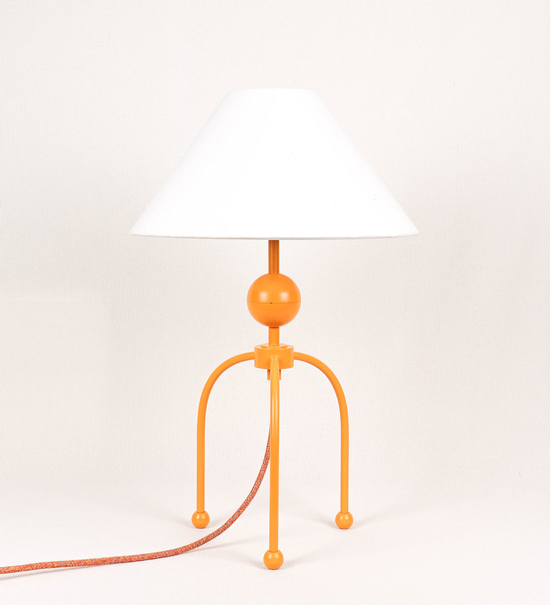 Candeeiro de mesa, de tom laranja-tangerina, com um design moderno e minimalista. O candeeiro tem o corpo com três pés curvados, terminando em pequenas esferas. A estrutura dos pés é interligada por uma peça central esférica. O cabo é revestido têxtil com um padrão de linhas coloridas, com interruptor e ficha transparentes. O abatjour é de forma cónica, em algodão, de tom bege claro com pequenas pintas escuras, semelhante à baunilha.