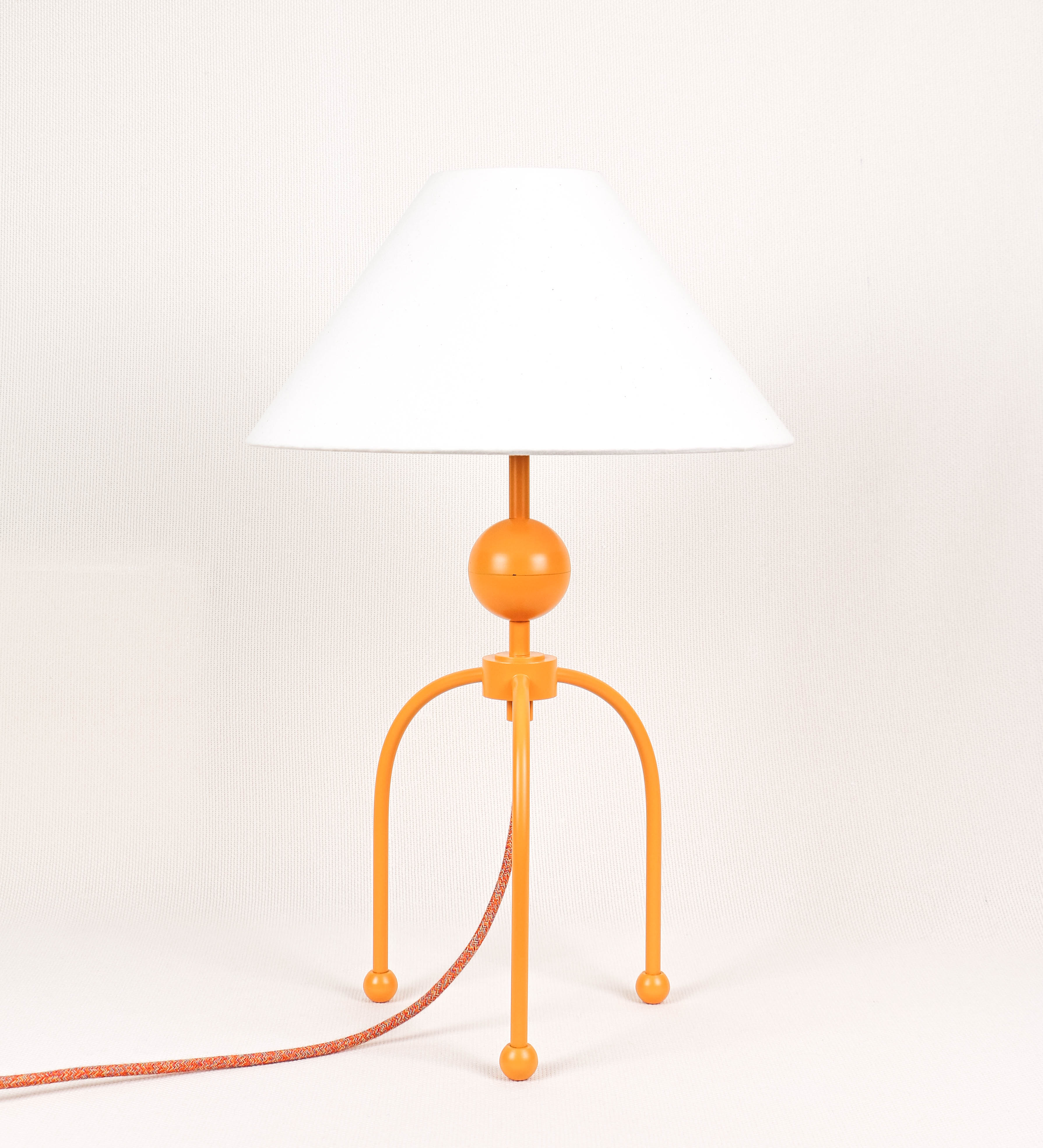 Candeeiro de mesa, de tom laranja-tangerina, com um design moderno e minimalista. O candeeiro tem o corpo com três pés curvados, terminando em pequenas esferas. A estrutura dos pés é interligada por uma peça central esférica. O cabo é revestido têxtil com um padrão de linhas coloridas, com interruptor e ficha transparentes. O abatjour é de forma cónica, em algodão, de tom bege claro com pequenas pintas escuras, semelhante à baunilha.