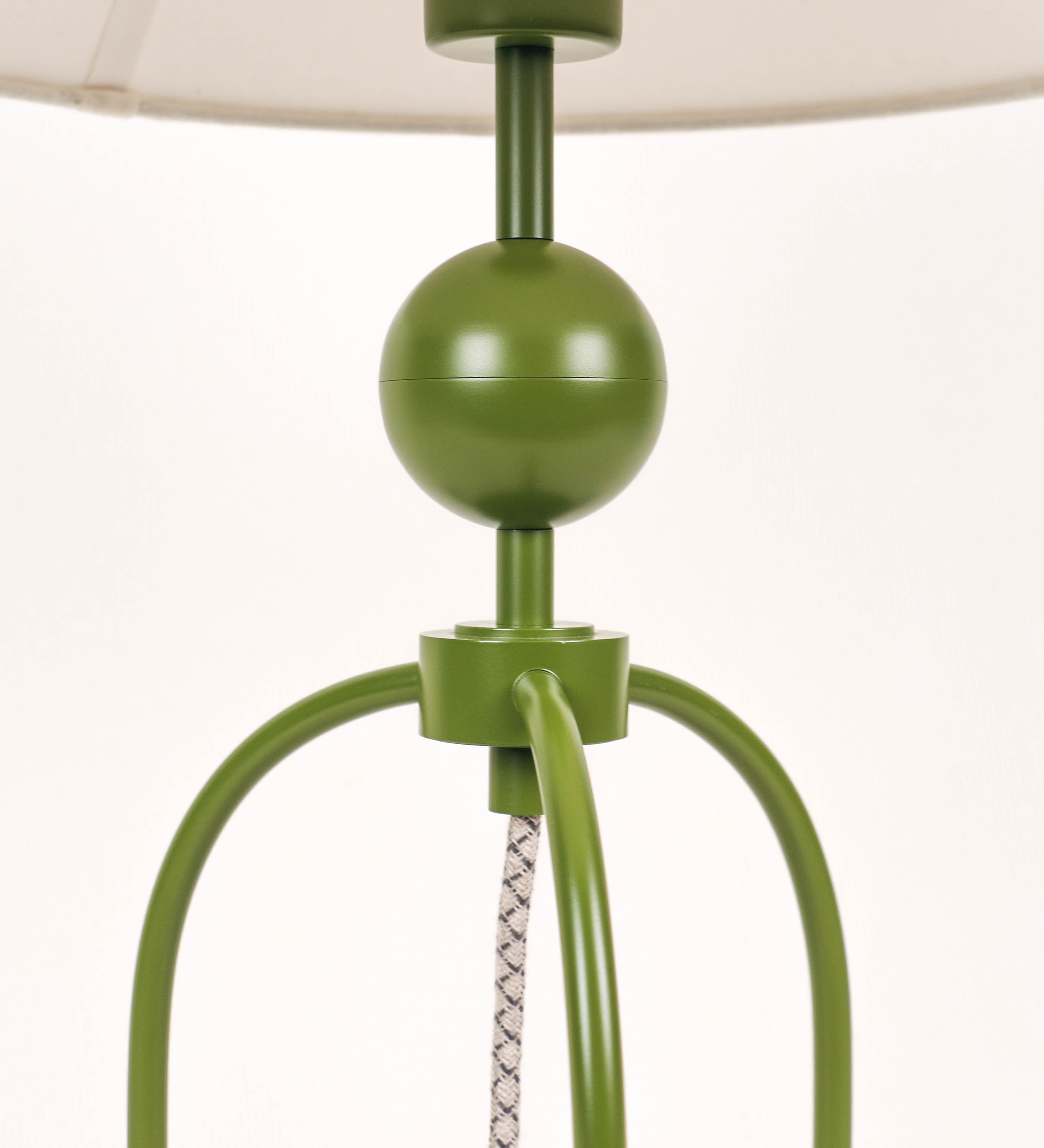 Candeeiro de mesa, de tom verde couve-de-bruxelas, com um design moderno e minimalista. O candeeiro tem o corpo com três pés curvados, terminando em pequenas esferas. A estrutura dos pés é interligada por uma peça central esférica. O cabo é revestido têxtil com um padrão de linhas castanhas e beges entrelaçadas, com interruptor e ficha transparentes. O abatjour é de forma cónica, em algodão, de tom bege claro com pequenas pintas escuras, semelhante à baunilha.