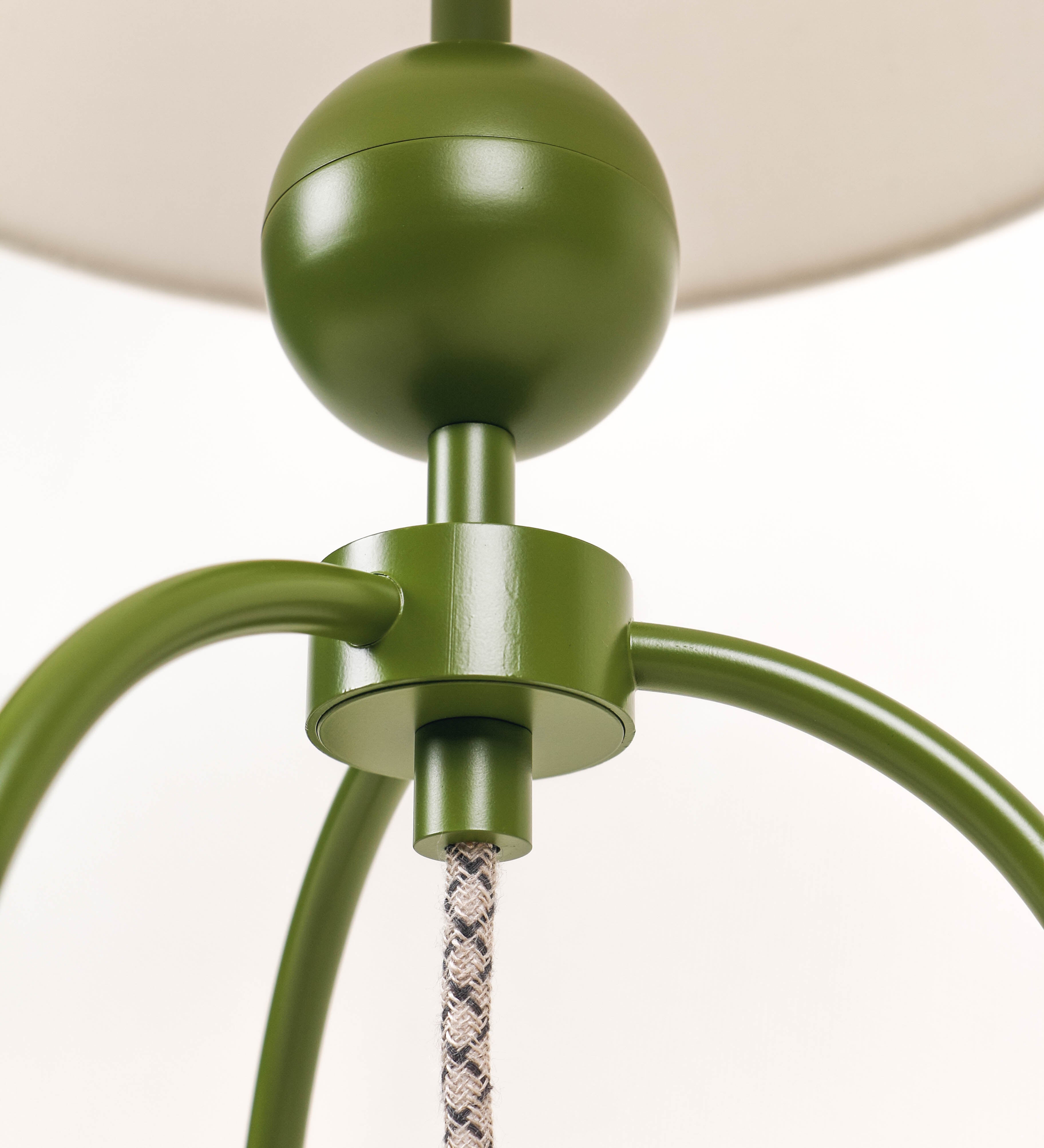 Candeeiro de mesa, de tom verde couve-de-bruxelas, com um design moderno e minimalista. O candeeiro tem o corpo com três pés curvados, terminando em pequenas esferas. A estrutura dos pés é interligada por uma peça central esférica. O cabo é revestido têxtil com um padrão de linhas castanhas e beges entrelaçadas, com interruptor e ficha transparentes. O abatjour é de forma cónica, em algodão, de tom bege claro com pequenas pintas escuras, semelhante à baunilha.