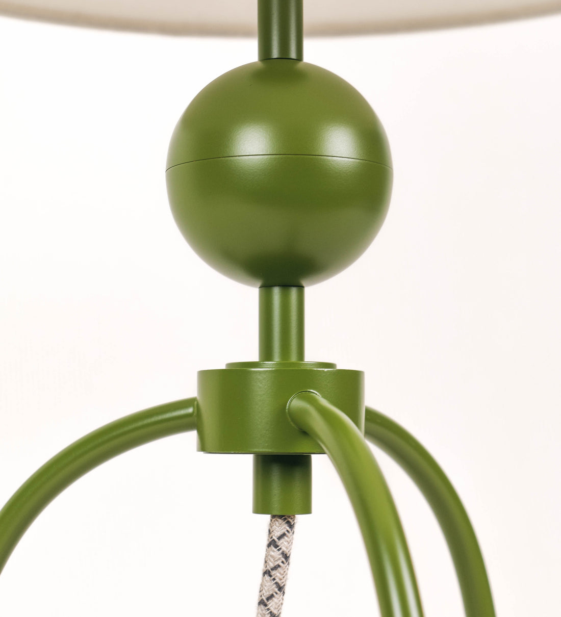 Candeeiro de mesa, de tom verde couve-de-bruxelas, com um design moderno e minimalista. O candeeiro tem o corpo com três pés curvados, terminando em pequenas esferas. A estrutura dos pés é interligada por uma peça central esférica. O cabo é revestido têxtil com um padrão de linhas castanhas e beges entrelaçadas, com interruptor e ficha transparentes. O abatjour é de forma cónica, em algodão, de tom bege claro com pequenas pintas escuras, semelhante à baunilha.