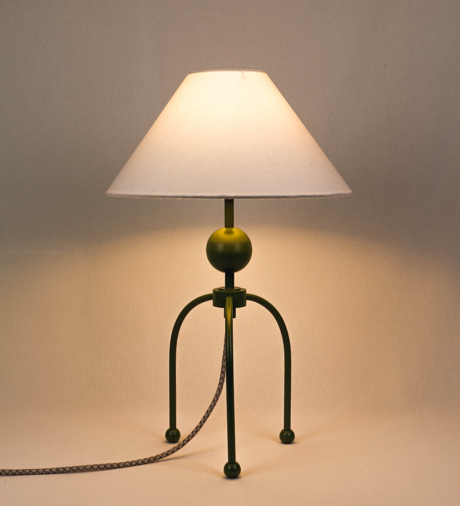 Candeeiro de mesa, de tom verde couve-de-bruxelas, com um design moderno e minimalista. O candeeiro tem o corpo com três pés curvados, terminando em pequenas esferas. A estrutura dos pés é interligada por uma peça central esférica. O cabo é revestido têxtil com um padrão de linhas castanhas e beges entrelaçadas, com interruptor e ficha transparentes. O abatjour é de forma cónica, em algodão, de tom bege claro com pequenas pintas escuras, semelhante à baunilha.