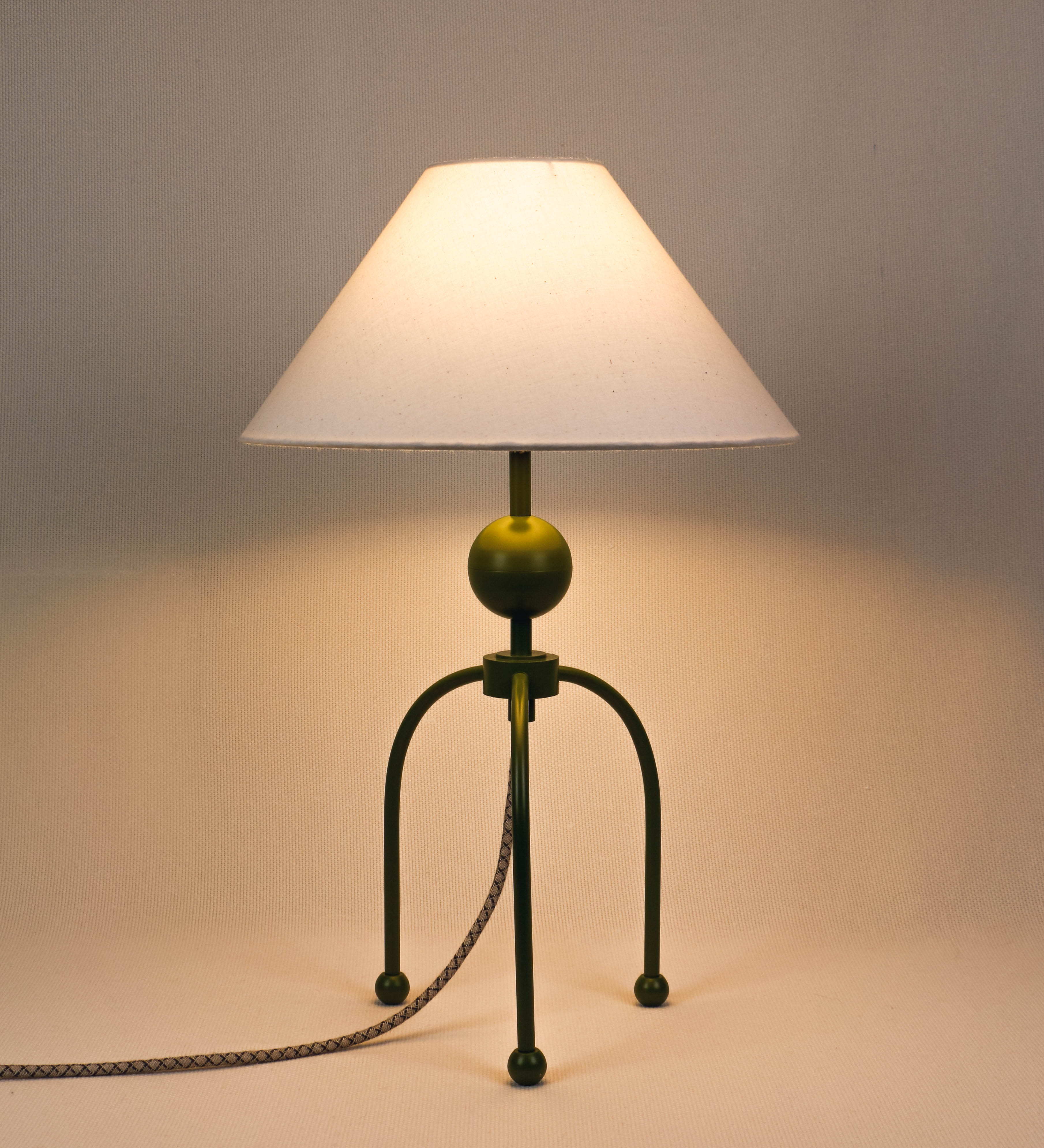 Candeeiro de mesa, de tom verde couve-de-bruxelas, com um design moderno e minimalista. O candeeiro tem o corpo com três pés curvados, terminando em pequenas esferas. A estrutura dos pés é interligada por uma peça central esférica. O cabo é revestido têxtil com um padrão de linhas castanhas e beges entrelaçadas, com interruptor e ficha transparentes. O abatjour é de forma cónica, em algodão, de tom bege claro com pequenas pintas escuras, semelhante à baunilha.