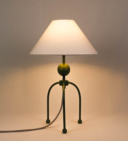 Candeeiro de mesa, de tom verde couve-de-bruxelas, com um design moderno e minimalista. O candeeiro tem o corpo com três pés curvados, terminando em pequenas esferas. A estrutura dos pés é interligada por uma peça central esférica. O cabo é revestido têxtil com um padrão de linhas castanhas e beges entrelaçadas, com interruptor e ficha transparentes. O abatjour é de forma cónica, em algodão, de tom bege claro com pequenas pintas escuras, semelhante à baunilha.