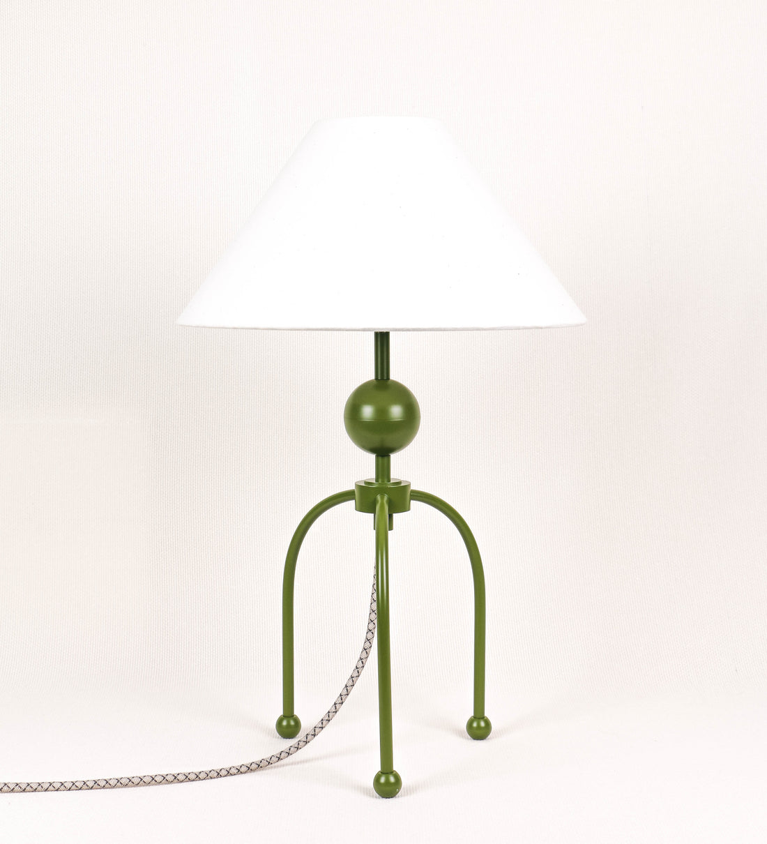 Candeeiro de mesa, de tom verde couve-de-bruxelas, com um design moderno e minimalista. O candeeiro tem o corpo com três pés curvados, terminando em pequenas esferas. A estrutura dos pés é interligada por uma peça central esférica. O cabo é revestido têxtil com um padrão de linhas castanhas e beges entrelaçadas, com interruptor e ficha transparentes. O abatjour é de forma cónica, em algodão, de tom bege claro com pequenas pintas escuras, semelhante à baunilha.