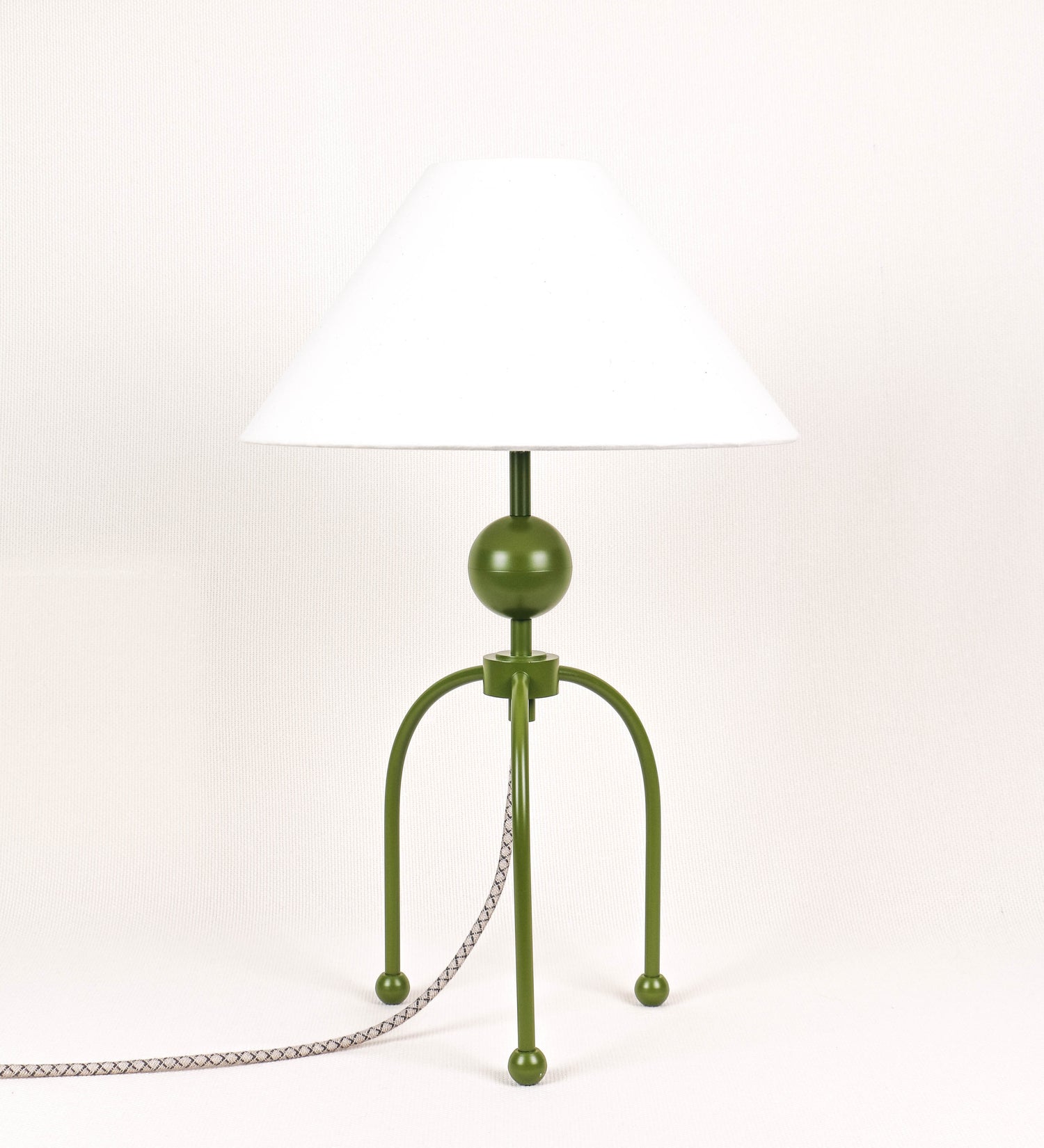 Candeeiro de mesa, de tom verde couve-de-bruxelas, com um design moderno e minimalista. O candeeiro tem o corpo com três pés curvados, terminando em pequenas esferas. A estrutura dos pés é interligada por uma peça central esférica. O cabo é revestido têxtil com um padrão de linhas castanhas e beges entrelaçadas, com interruptor e ficha transparentes. O abatjour é de forma cónica, em algodão, de tom bege claro com pequenas pintas escuras, semelhante à baunilha.