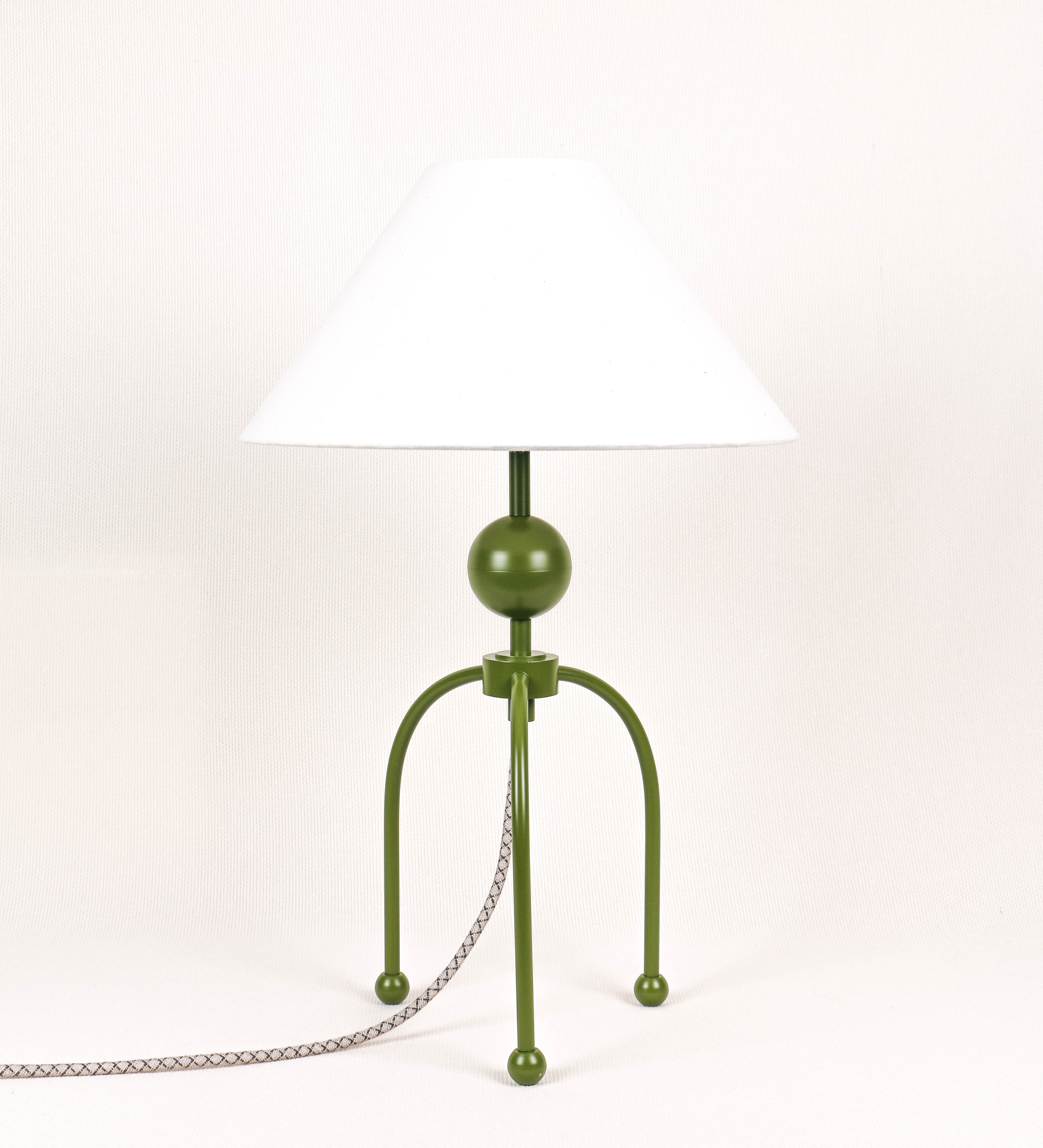 Candeeiro de mesa, de tom verde couve-de-bruxelas, com um design moderno e minimalista. O candeeiro tem o corpo com três pés curvados, terminando em pequenas esferas. A estrutura dos pés é interligada por uma peça central esférica. O cabo é revestido têxtil com um padrão de linhas castanhas e beges entrelaçadas, com interruptor e ficha transparentes. O abatjour é de forma cónica, em algodão, de tom bege claro com pequenas pintas escuras, semelhante à baunilha.