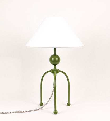 Candeeiro de mesa, de tom verde couve-de-bruxelas, com um design moderno e minimalista. O candeeiro tem o corpo com três pés curvados, terminando em pequenas esferas. A estrutura dos pés é interligada por uma peça central esférica. O cabo é revestido têxtil com um padrão de linhas castanhas e beges entrelaçadas, com interruptor e ficha transparentes. O abatjour é de forma cónica, em algodão, de tom bege claro com pequenas pintas escuras, semelhante à baunilha.