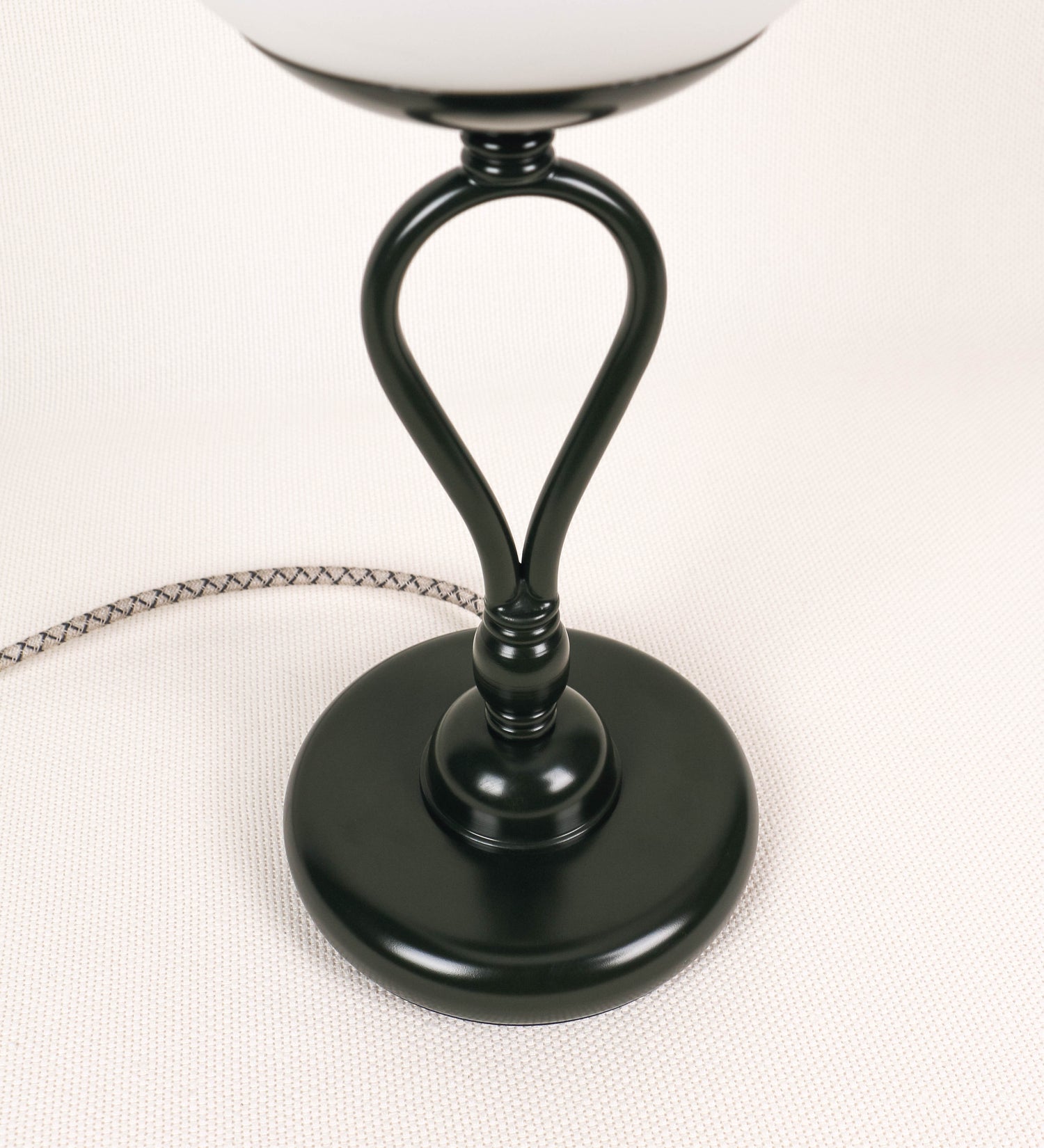 Candeeiro de mesa com um design clássico e sofisticado. O candeeiro possui uma base circular e robusta de tom verde-azeitona. A estrutura da base é complementada por um suporte em forma de nó, também de tom verde-azeitona, que se eleva graciosamente até ao suporte da luz. A luz é difundida através de uma esfera de vidro fosco branco, assente no corpo. O cabo é revestido têxtil com um padrão trançado com linhas castanhas e beges, com interruptor e ficha transparentes.