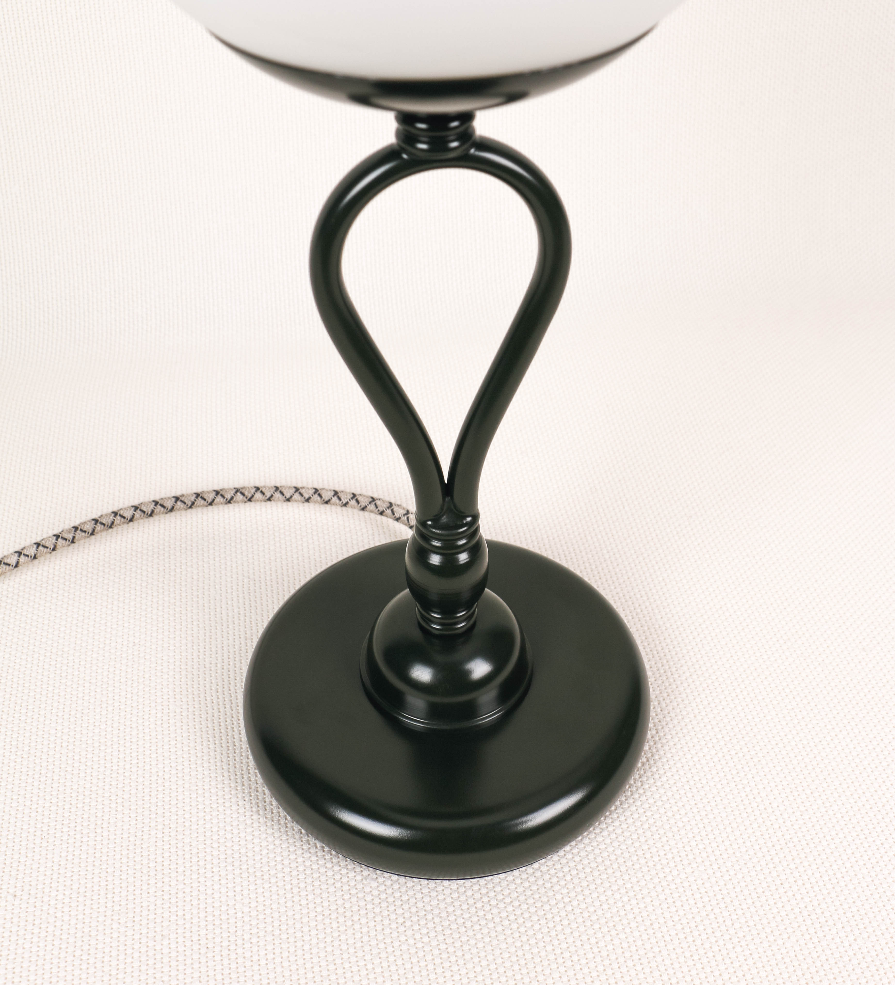Candeeiro de mesa com um design clássico e sofisticado. O candeeiro possui uma base circular e robusta de tom verde-azeitona. A estrutura da base é complementada por um suporte em forma de nó, também de tom verde-azeitona, que se eleva graciosamente até ao suporte da luz. A luz é difundida através de uma esfera de vidro fosco branco, assente no corpo. O cabo é revestido têxtil com um padrão trançado com linhas castanhas e beges, com interruptor e ficha transparentes.