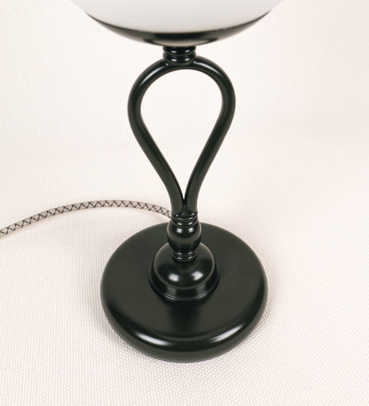 Candeeiro de mesa com um design clássico e sofisticado. O candeeiro possui uma base circular e robusta de tom verde-azeitona. A estrutura da base é complementada por um suporte em forma de nó, também de tom verde-azeitona, que se eleva graciosamente até ao suporte da luz. A luz é difundida através de uma esfera de vidro fosco branco, assente no corpo. O cabo é revestido têxtil com um padrão trançado com linhas castanhas e beges, com interruptor e ficha transparentes.