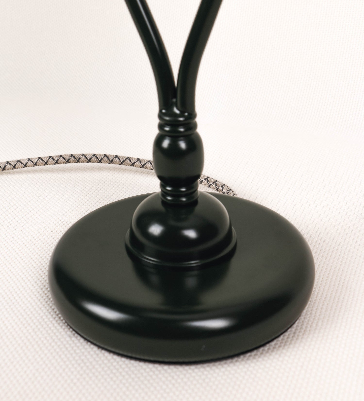 Candeeiro de mesa com um design clássico e sofisticado. O candeeiro possui uma base circular e robusta de tom verde-azeitona. A estrutura da base é complementada por um suporte em forma de nó, também de tom verde-azeitona, que se eleva graciosamente até ao suporte da luz. A luz é difundida através de uma esfera de vidro fosco branco, assente no corpo. O cabo é revestido têxtil com um padrão trançado com linhas castanhas e beges, com interruptor e ficha transparentes.