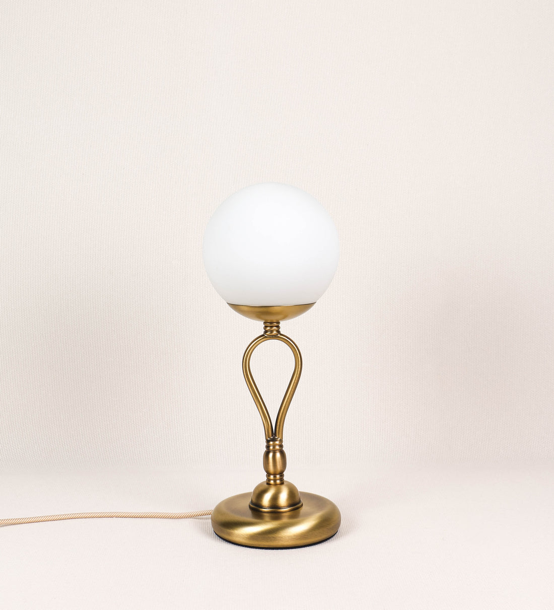 Candeeiro de mesa com um design clássico e sofisticado. O candeeiro possui uma base circular e robusta em latão com acabamento em ouro velho. A estrutura da base é complementada por um suporte em forma de nó, também em ouro velho, que se eleva graciosamente até ao suporte da luz. A luz é difundida através de uma esfera de vidro fosco branco, assente no corpo. O cabo é revestido têxtil com um padrão trançado de linhas douradas e brancas, com interruptor e ficha transparentes.