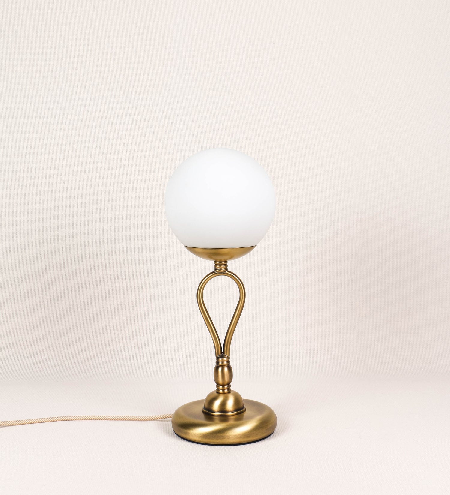 Candeeiro de mesa com um design clássico e sofisticado. O candeeiro possui uma base circular e robusta em latão com acabamento em ouro velho. A estrutura da base é complementada por um suporte em forma de nó, também em ouro velho, que se eleva graciosamente até ao suporte da luz. A luz é difundida através de uma esfera de vidro fosco branco, assente no corpo. O cabo é revestido têxtil com um padrão trançado de linhas douradas e brancas, com interruptor e ficha transparentes.