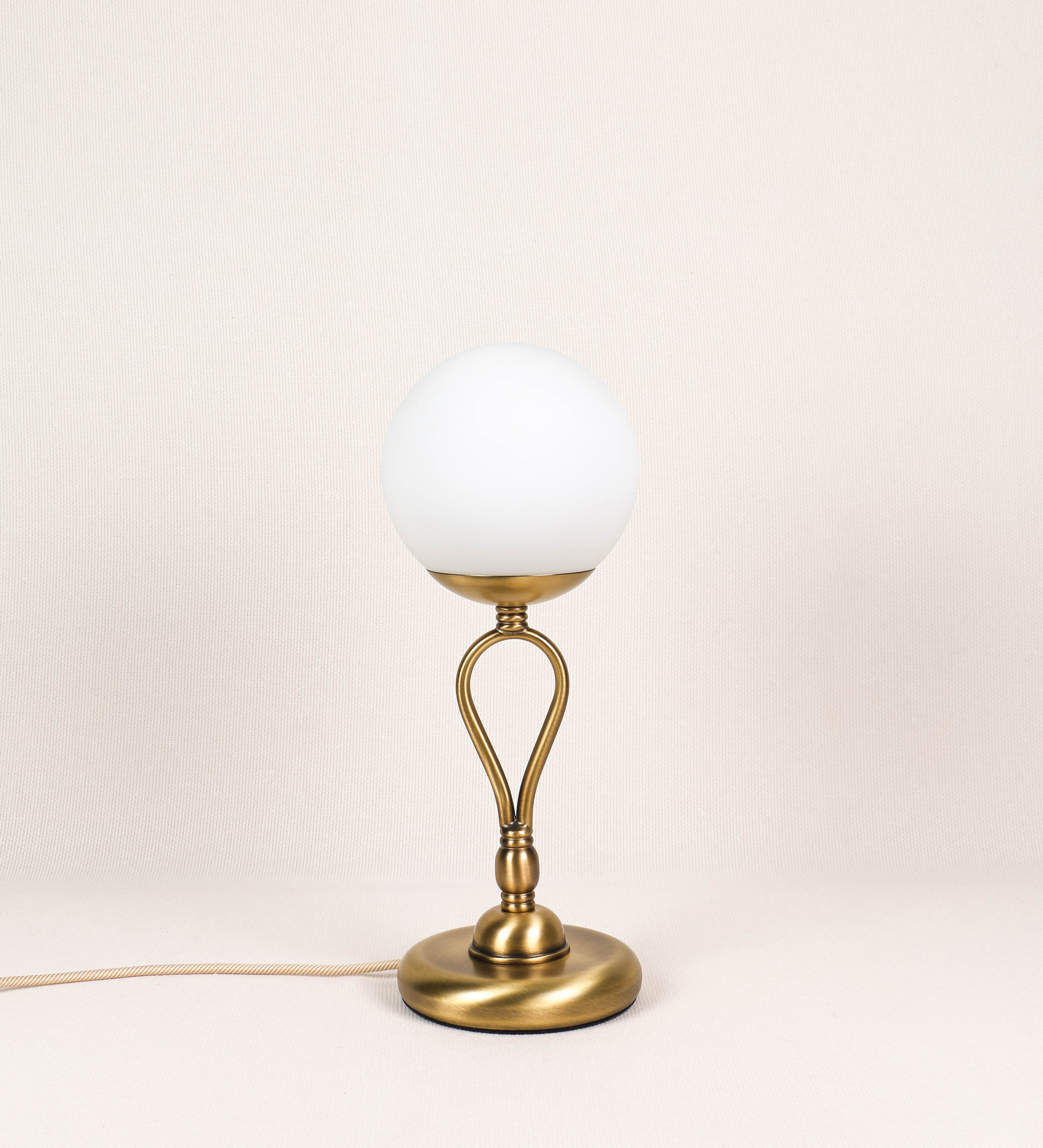 Candeeiro de mesa com um design clássico e sofisticado. O candeeiro possui uma base circular e robusta em latão com acabamento em ouro velho. A estrutura da base é complementada por um suporte em forma de nó, também em ouro velho, que se eleva graciosamente até ao suporte da luz. A luz é difundida através de uma esfera de vidro fosco branco, assente no corpo. O cabo é revestido têxtil com um padrão trançado de linhas douradas e brancas, com interruptor e ficha transparentes.