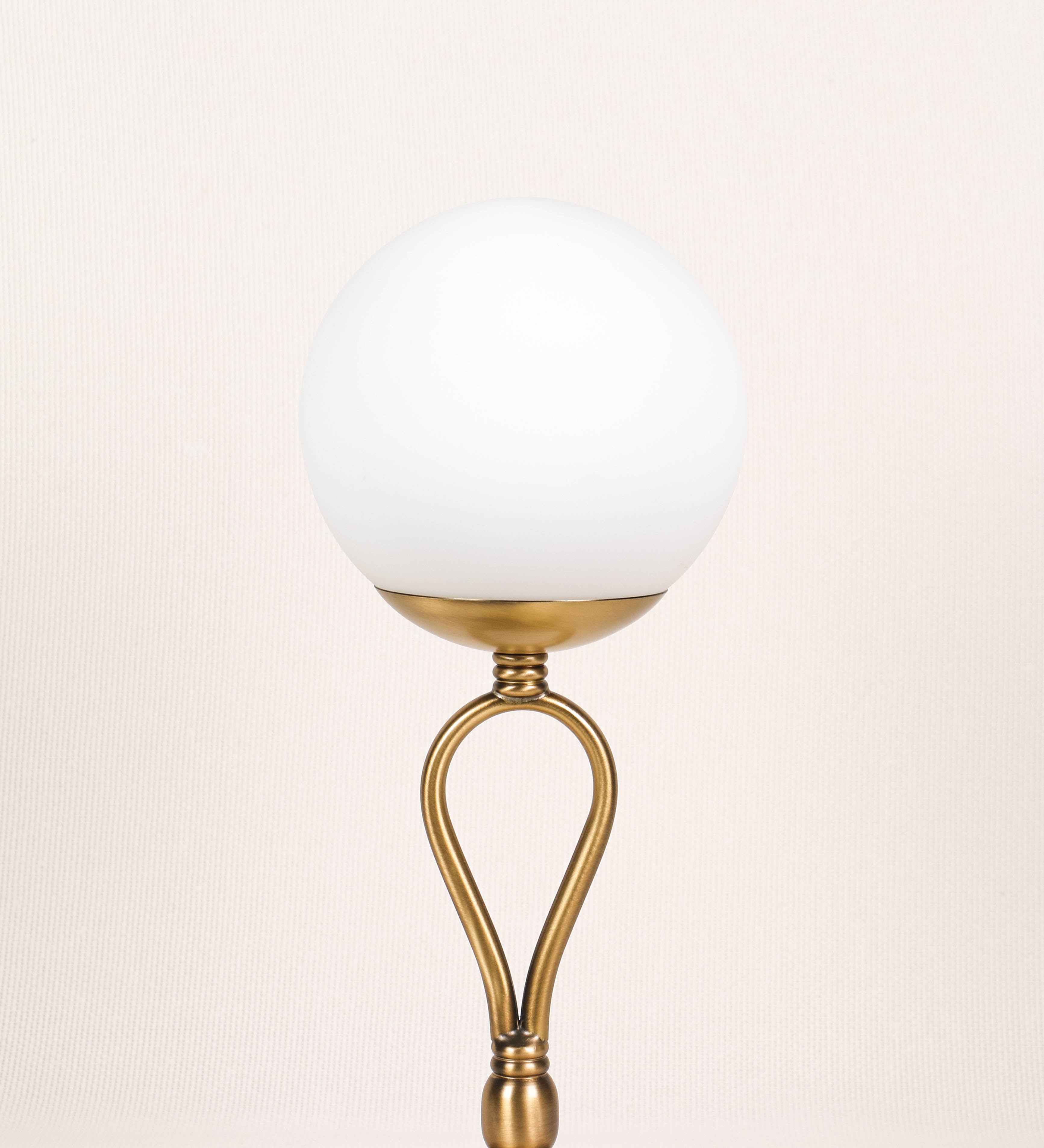 Candeeiro de mesa com um design clássico e sofisticado. O candeeiro possui uma base circular e robusta em latão com acabamento em ouro velho. A estrutura da base é complementada por um suporte em forma de nó, também em ouro velho, que se eleva graciosamente até ao suporte da luz. A luz é difundida através de uma esfera de vidro fosco branco, assente no corpo. O cabo é revestido têxtil com um padrão trançado de linhas douradas e brancas, com interruptor e ficha transparentes.