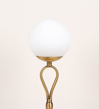 Candeeiro de mesa com um design clássico e sofisticado. O candeeiro possui uma base circular e robusta em latão com acabamento em ouro velho. A estrutura da base é complementada por um suporte em forma de nó, também em ouro velho, que se eleva graciosamente até ao suporte da luz. A luz é difundida através de uma esfera de vidro fosco branco, assente no corpo. O cabo é revestido têxtil com um padrão trançado de linhas douradas e brancas, com interruptor e ficha transparentes.