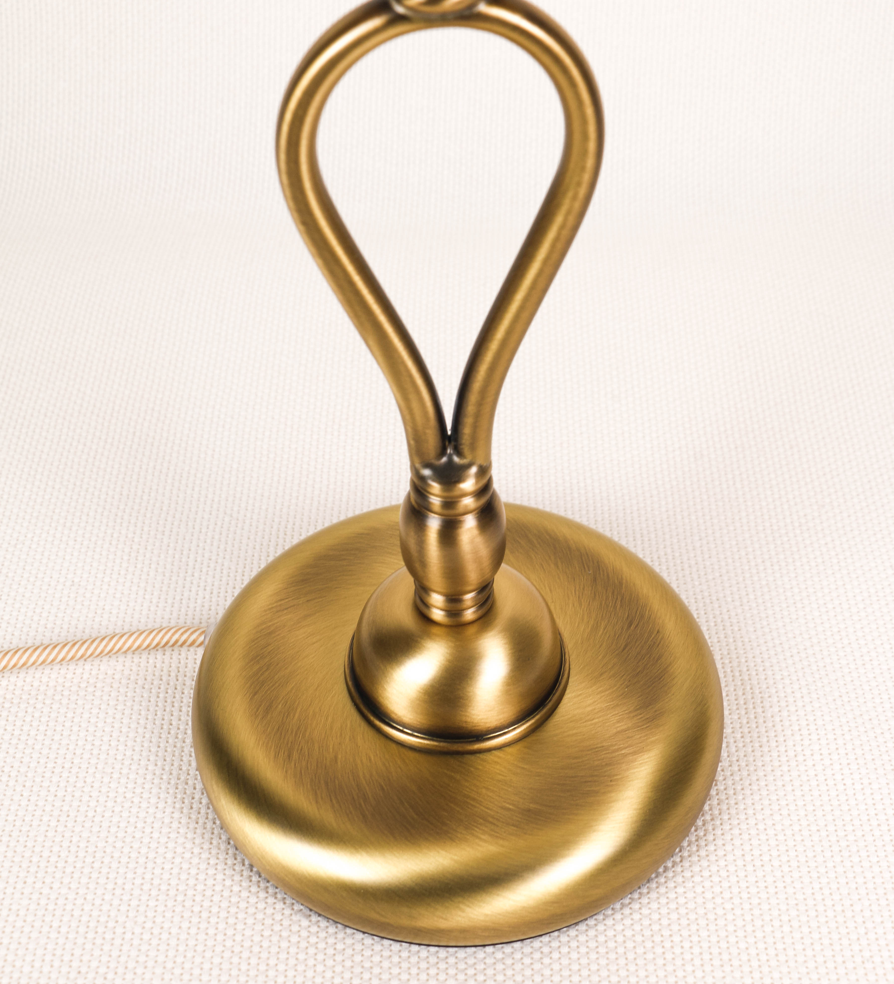 Candeeiro de mesa com um design clássico e sofisticado. O candeeiro possui uma base circular e robusta em latão com acabamento em ouro velho. A estrutura da base é complementada por um suporte em forma de nó, também em ouro velho, que se eleva graciosamente até ao suporte da luz. A luz é difundida através de uma esfera de vidro fosco branco, assente no corpo. O cabo é revestido têxtil com um padrão trançado de linhas douradas e brancas, com interruptor e ficha transparentes.