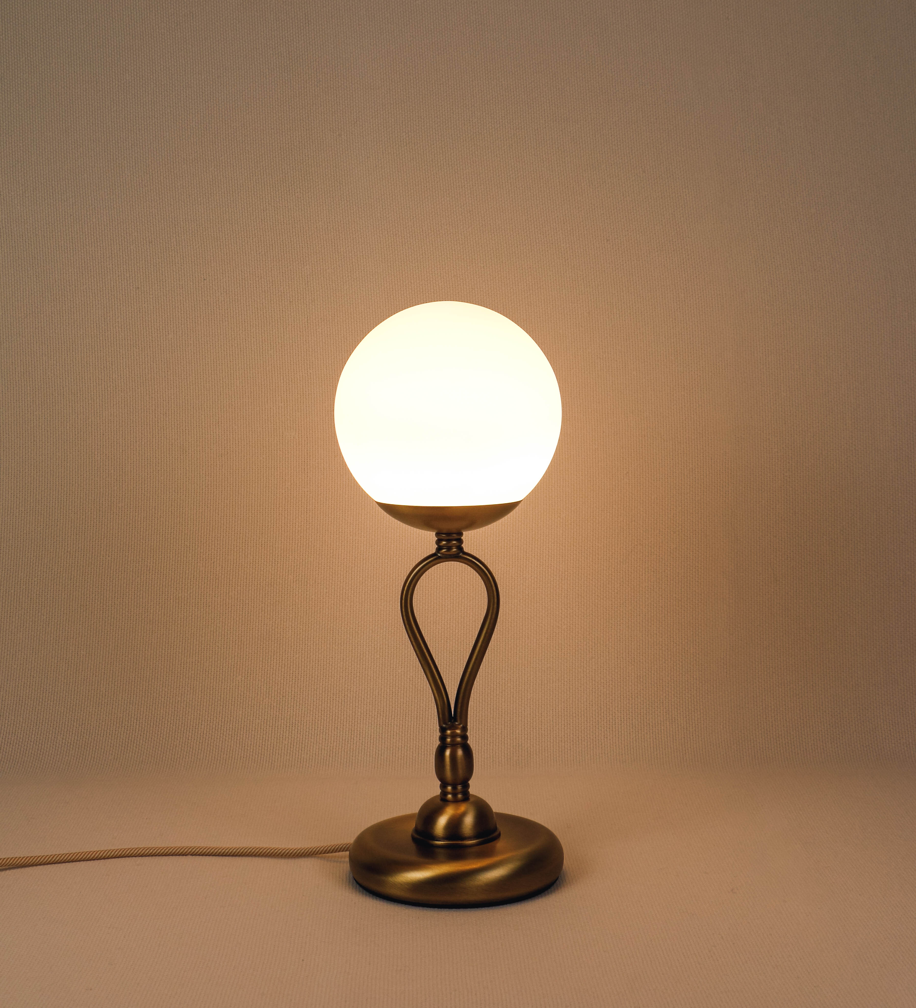 Candeeiro de mesa com um design clássico e sofisticado. O candeeiro possui uma base circular e robusta em latão com acabamento em ouro velho. A estrutura da base é complementada por um suporte em forma de nó, também em ouro velho, que se eleva graciosamente até ao suporte da luz. A luz é difundida através de uma esfera de vidro fosco branco, assente no corpo. O cabo é revestido têxtil com um padrão trançado de linhas douradas e brancas, com interruptor e ficha transparentes.