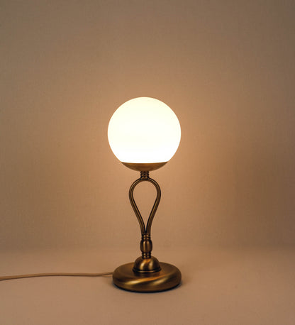 Candeeiro de mesa com um design clássico e sofisticado. O candeeiro possui uma base circular e robusta em latão com acabamento em ouro velho. A estrutura da base é complementada por um suporte em forma de nó, também em ouro velho, que se eleva graciosamente até ao suporte da luz. A luz é difundida através de uma esfera de vidro fosco branco, assente no corpo. O cabo é revestido têxtil com um padrão trançado de linhas douradas e brancas, com interruptor e ficha transparentes.