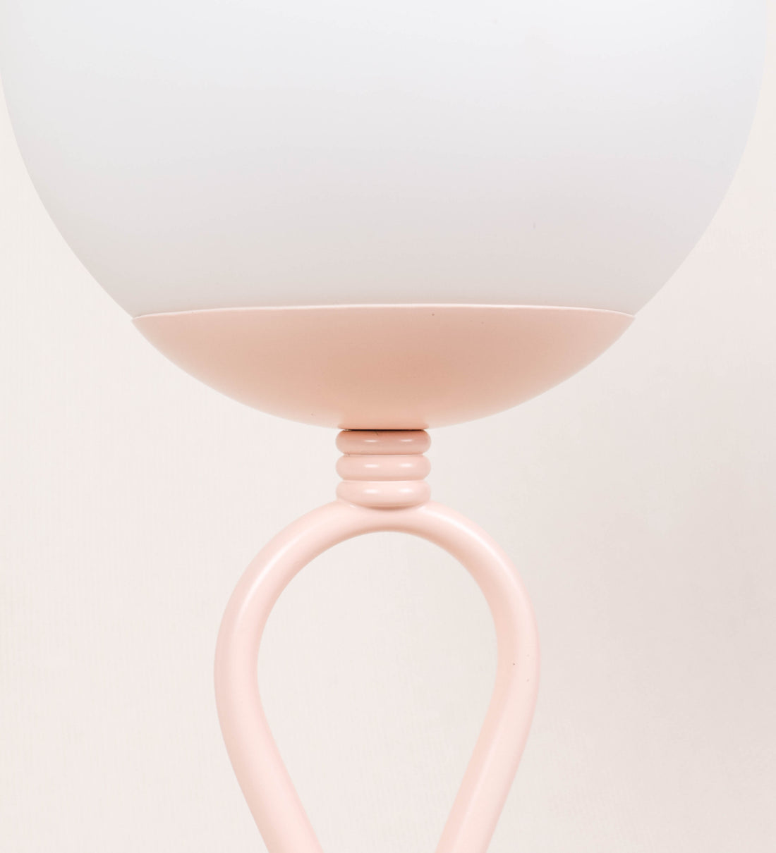 Candeeiro de mesa com um design clássico e sofisticado. O candeeiro possui uma base circular e robusta de tom rosa claro. A estrutura da base é complementada por um suporte em forma de nó, também de tom rosa claro, que se eleva graciosamente até ao suporte da luz. A luz é difundida através de uma esfera de vidro fosco branco, assente no corpo. O cabo é revestido têxtil com um padrão trançado de tom rosa velho, com interruptor e ficha transparentes.