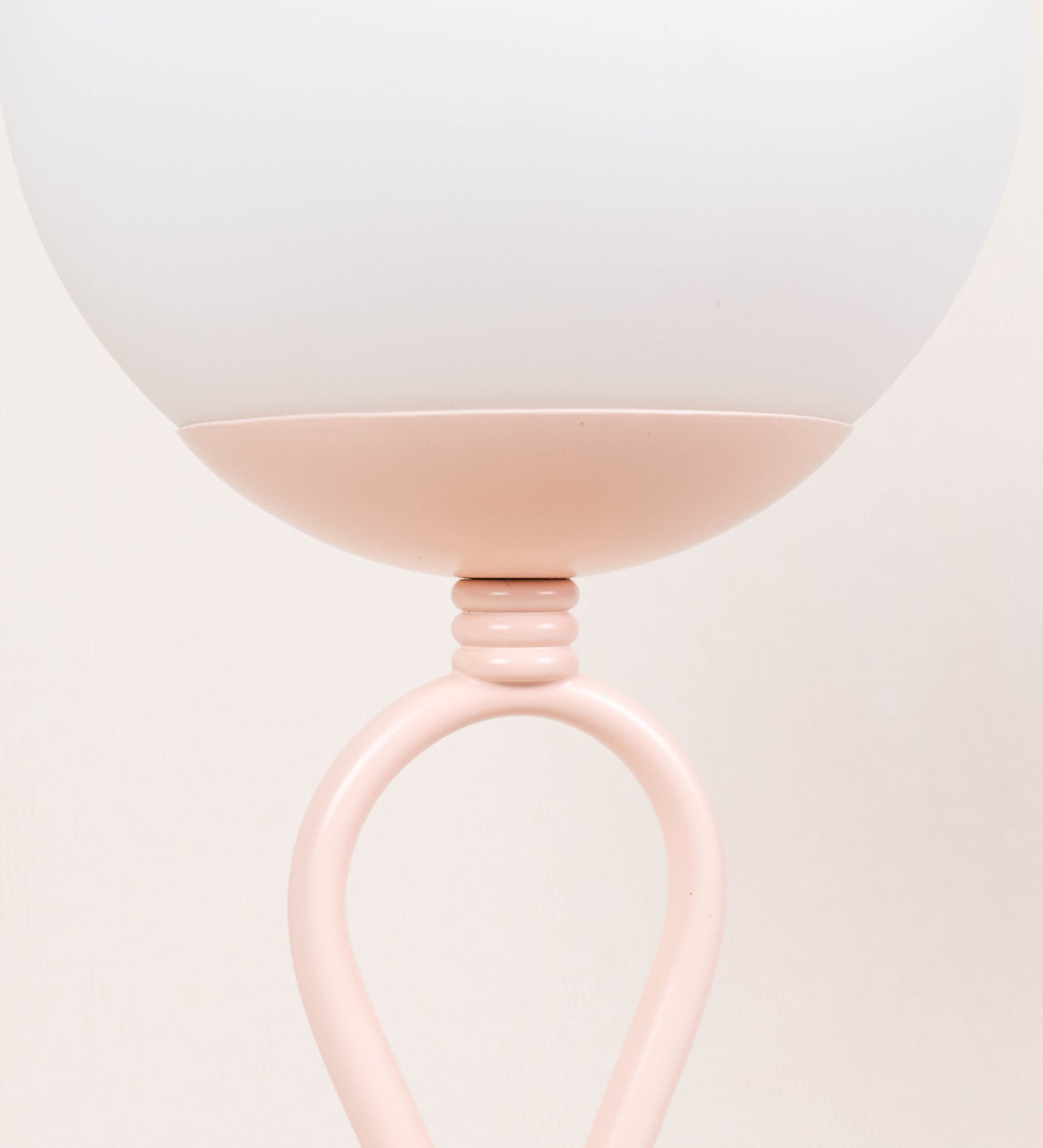 Candeeiro de mesa com um design clássico e sofisticado. O candeeiro possui uma base circular e robusta de tom rosa claro. A estrutura da base é complementada por um suporte em forma de nó, também de tom rosa claro, que se eleva graciosamente até ao suporte da luz. A luz é difundida através de uma esfera de vidro fosco branco, assente no corpo. O cabo é revestido têxtil com um padrão trançado de tom rosa velho, com interruptor e ficha transparentes.