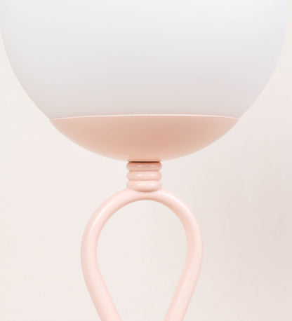 Candeeiro de mesa com um design clássico e sofisticado. O candeeiro possui uma base circular e robusta de tom rosa claro. A estrutura da base é complementada por um suporte em forma de nó, também de tom rosa claro, que se eleva graciosamente até ao suporte da luz. A luz é difundida através de uma esfera de vidro fosco branco, assente no corpo. O cabo é revestido têxtil com um padrão trançado de tom rosa velho, com interruptor e ficha transparentes.