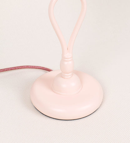 Candeeiro de mesa com um design clássico e sofisticado. O candeeiro possui uma base circular e robusta de tom rosa claro. A estrutura da base é complementada por um suporte em forma de nó, também de tom rosa claro, que se eleva graciosamente até ao suporte da luz. A luz é difundida através de uma esfera de vidro fosco branco, assente no corpo. O cabo é revestido têxtil com um padrão trançado de tom rosa velho, com interruptor e ficha transparentes.