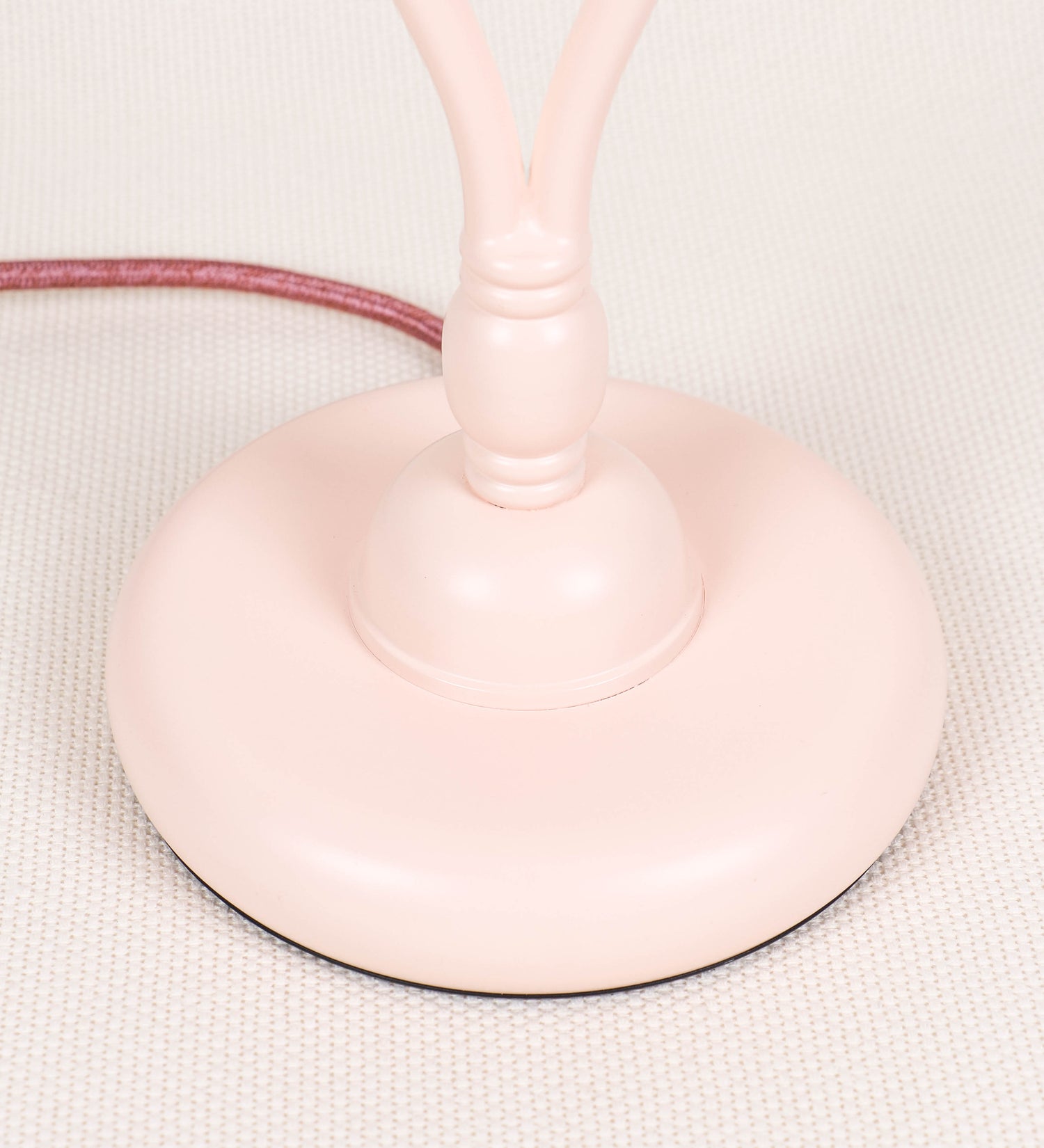 Candeeiro de mesa com um design clássico e sofisticado. O candeeiro possui uma base circular e robusta de tom rosa claro. A estrutura da base é complementada por um suporte em forma de nó, também de tom rosa claro, que se eleva graciosamente até ao suporte da luz. A luz é difundida através de uma esfera de vidro fosco branco, assente no corpo. O cabo é revestido têxtil com um padrão trançado de tom rosa velho, com interruptor e ficha transparentes.