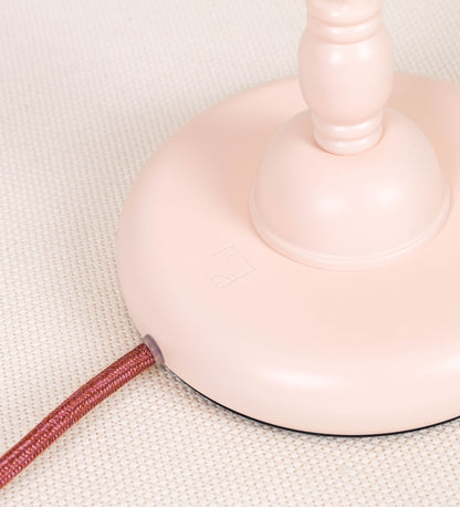 Candeeiro de mesa com um design clássico e sofisticado. O candeeiro possui uma base circular e robusta de tom rosa claro. A estrutura da base é complementada por um suporte em forma de nó, também de tom rosa claro, que se eleva graciosamente até ao suporte da luz. A luz é difundida através de uma esfera de vidro fosco branco, assente no corpo. O cabo é revestido têxtil com um padrão trançado de tom rosa velho, com interruptor e ficha transparentes.