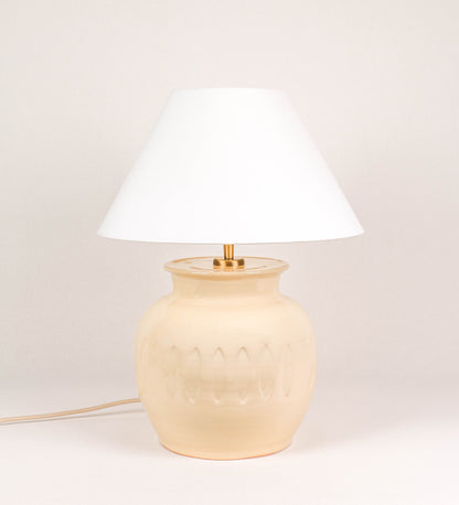 Candeeiro de mesa com um design rústico e sofisticado. A base do candeeiro é de barro vidrado de tom bege claro. A forma da base é arredondada com uma borda superior mais estreita, lembrando um vaso tradicional. A superfície da base é decorada com um padrão de relevos ovais. O suporte da lâmpada é em ouro velho e é manipulável. O cabo é revestido têxtil com um padrão trançado de linhas douradas e brancas, com interruptor e ficha transparentes. O abatjour é cónico, em algodão, de tom bege claro.