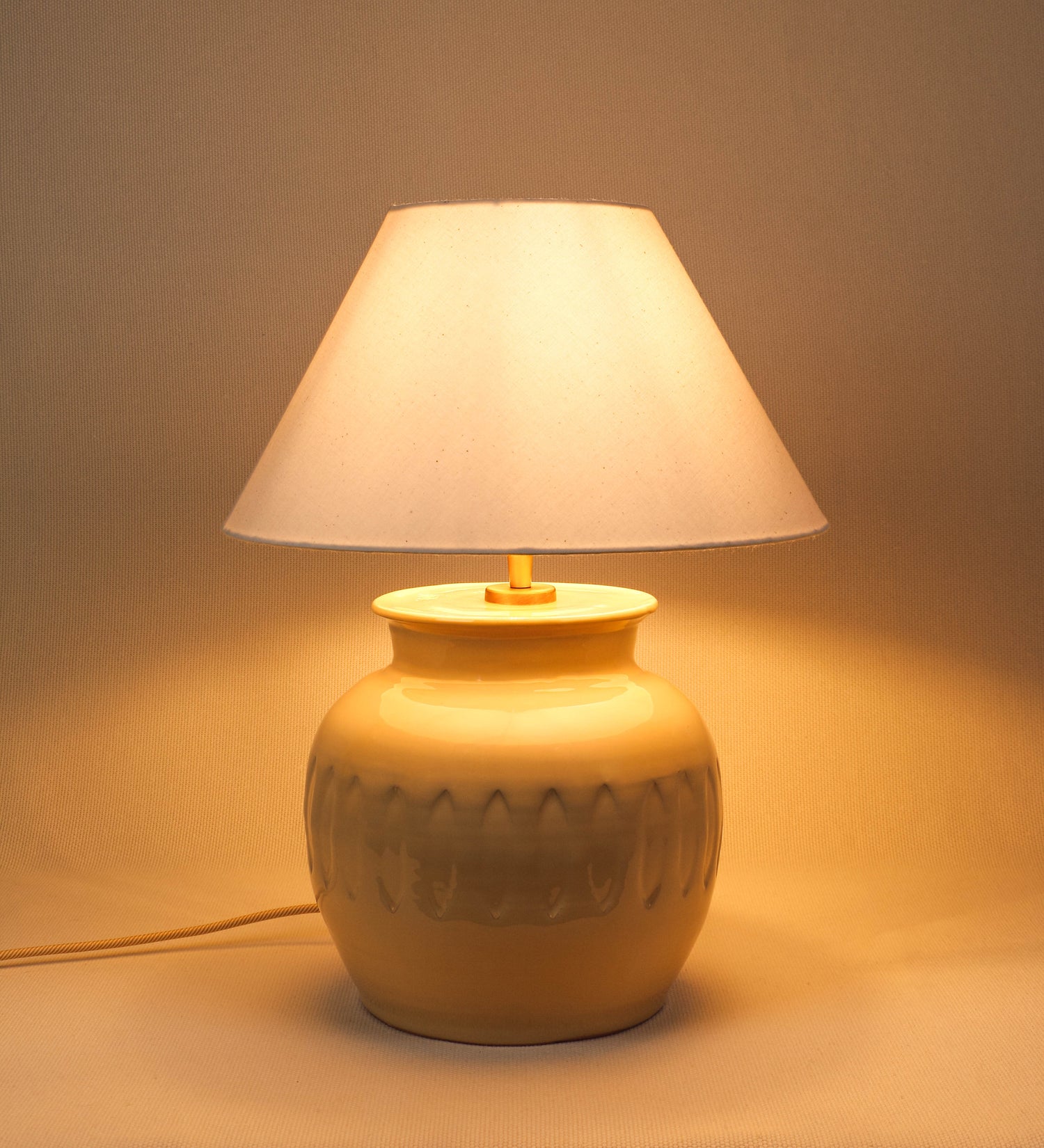 Candeeiro de mesa com um design rústico e sofisticado. A base do candeeiro é de barro vidrado de tom bege claro. A forma da base é arredondada com uma borda superior mais estreita, lembrando um vaso tradicional. A superfície da base é decorada com um padrão de relevos ovais. O suporte da lâmpada é em ouro velho e é manipulável. O cabo é revestido têxtil com um padrão trançado de linhas douradas e brancas, com interruptor e ficha transparentes. O abatjour é cónico, em algodão, de tom bege claro.