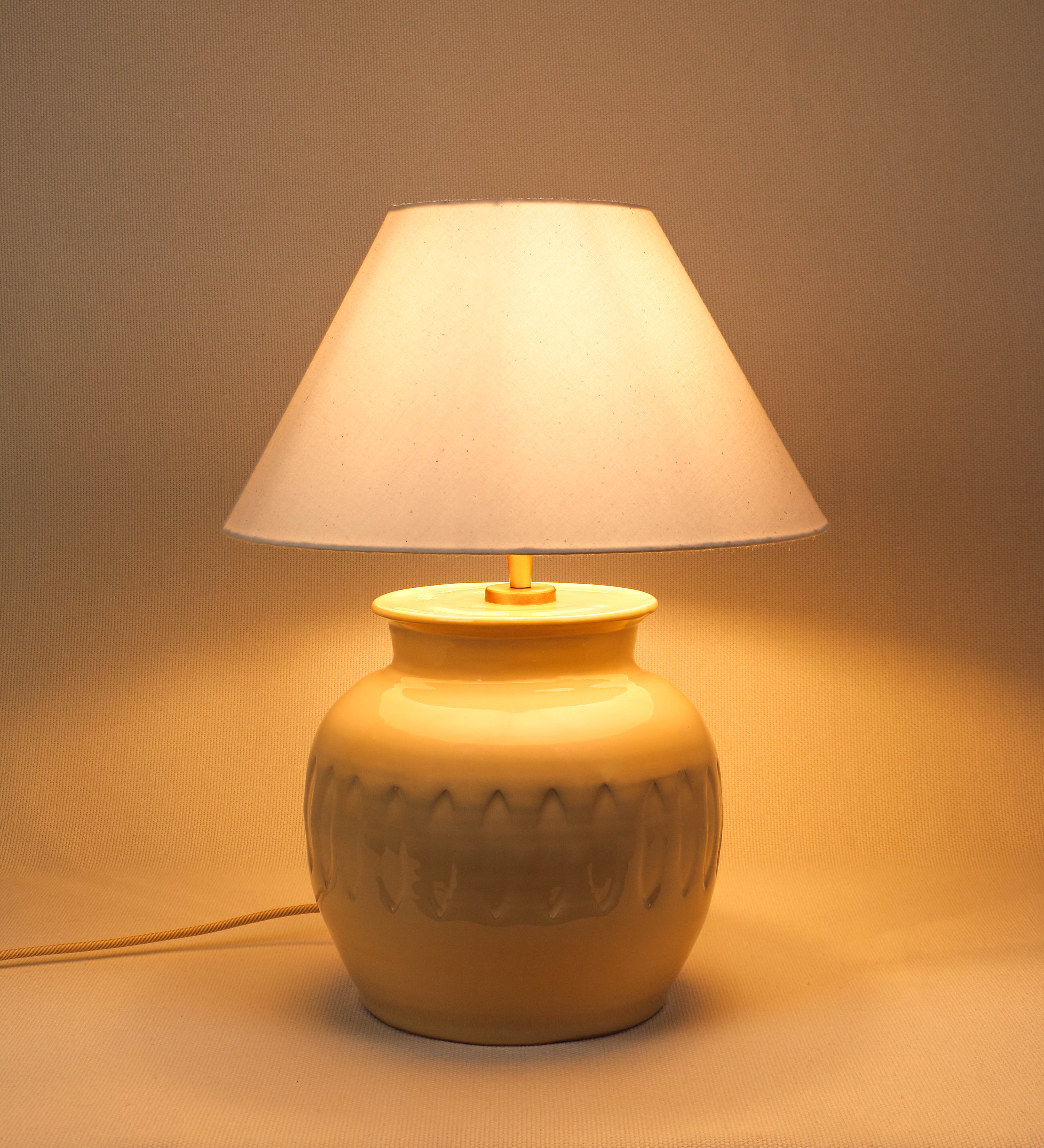 Candeeiro de mesa com um design rústico e sofisticado. A base do candeeiro é de barro vidrado de tom bege claro. A forma da base é arredondada com uma borda superior mais estreita, lembrando um vaso tradicional. A superfície da base é decorada com um padrão de relevos ovais. O suporte da lâmpada é em ouro velho e é manipulável. O cabo é revestido têxtil com um padrão trançado de linhas douradas e brancas, com interruptor e ficha transparentes. O abatjour é cónico, em algodão, de tom bege claro.