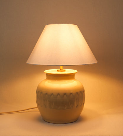 Candeeiro de mesa com um design rústico e sofisticado. A base do candeeiro é de barro vidrado de tom bege claro. A forma da base é arredondada com uma borda superior mais estreita, lembrando um vaso tradicional. A superfície da base é decorada com um padrão de relevos ovais. O suporte da lâmpada é em ouro velho e é manipulável. O cabo é revestido têxtil com um padrão trançado de linhas douradas e brancas, com interruptor e ficha transparentes. O abatjour é cónico, em algodão, de tom bege claro.