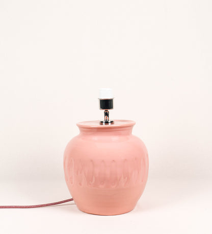 Candeeiro de mesa com um design rústico e sofisticado. A base do candeeiro é de barro vidrado de tom rosa salmão. A forma da base é arredondada com uma borda superior mais estreita, lembrando um vaso tradicional. A superfície da base é decorada com um padrão de relevos ovais. O suporte da lâmpada é em níquel polido e é manipulável. O cabo é revestido têxtil com um padrão trançado de tom rosa velho, com interruptor e ficha transparentes.