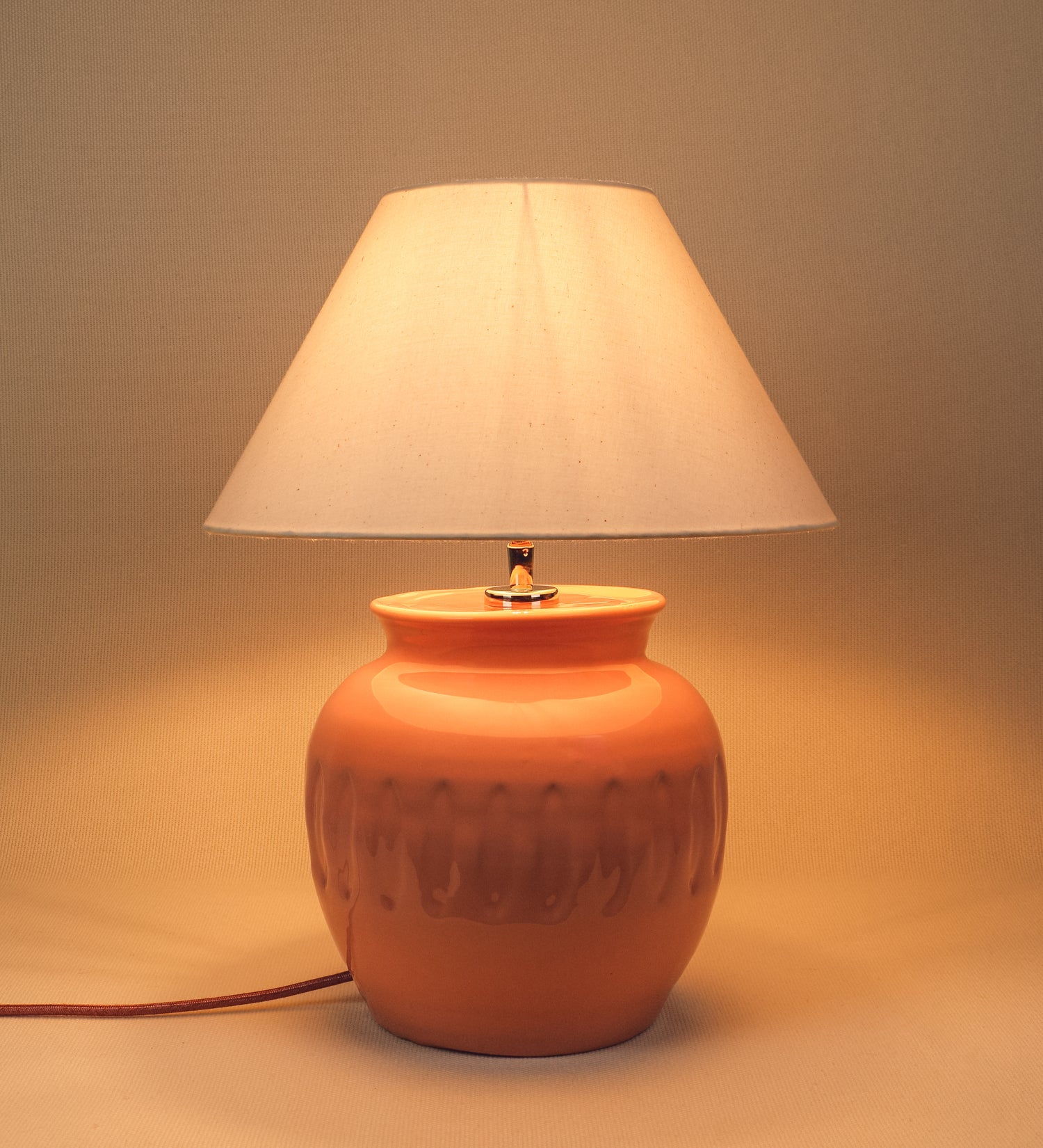 Candeeiro de mesa com um design rústico e sofisticado. A base do candeeiro é de barro vidrado de tom rosa salmão. A forma da base é arredondada com uma borda superior mais estreita, lembrando um vaso tradicional. A superfície da base é decorada com um padrão de relevos ovais. O suporte da lâmpada é em níquel polido e é manipulável. O cabo é revestido têxtil com um padrão trançado de tom rosa velho, com interruptor e ficha transparentes. O abatjour é cónico, em algodão, de tom bege claro.