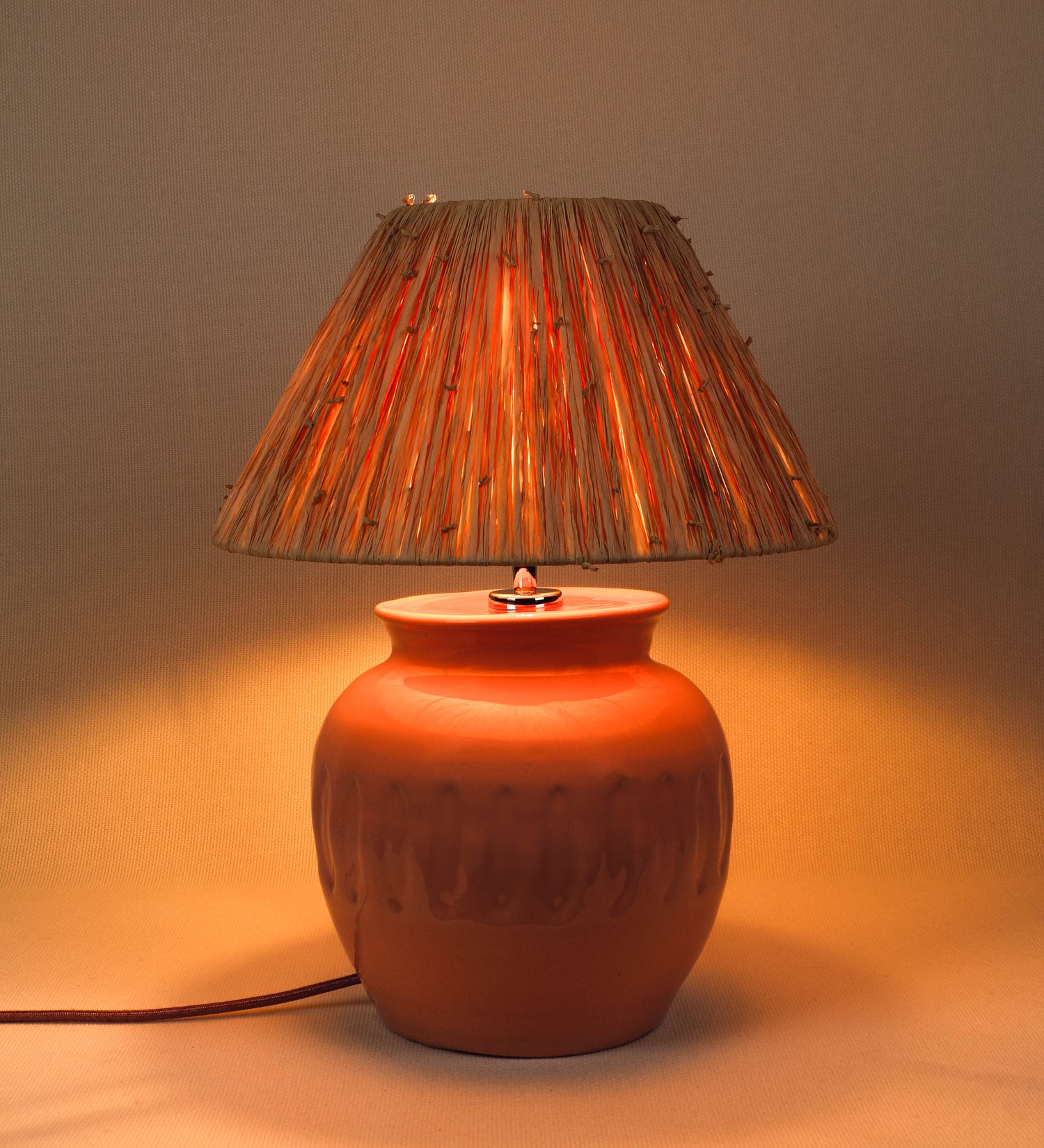 Candeeiro de mesa com um design rústico e sofisticado. A base do candeeiro é de barro vidrado de tom rosa salmão. A forma da base é arredondada com uma borda superior mais estreita, lembrando um vaso tradicional. A superfície da base é decorada com um padrão de relevos ovais. O suporte da lâmpada é em níquel polido e é manipulável. O cabo é revestido têxtil com um padrão trançado de tom rosa velho, com interruptor e ficha transparentes. O abatjour é cónico, de ráfia com nós.