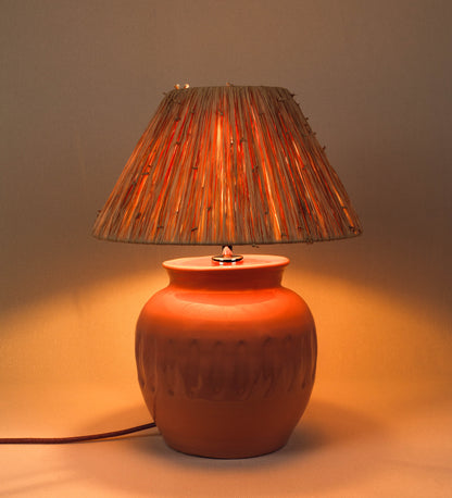 Candeeiro de mesa com um design rústico e sofisticado. A base do candeeiro é de barro vidrado de tom rosa salmão. A forma da base é arredondada com uma borda superior mais estreita, lembrando um vaso tradicional. A superfície da base é decorada com um padrão de relevos ovais. O suporte da lâmpada é em níquel polido e é manipulável. O cabo é revestido têxtil com um padrão trançado de tom rosa velho, com interruptor e ficha transparentes. O abatjour é cónico, de ráfia com nós.