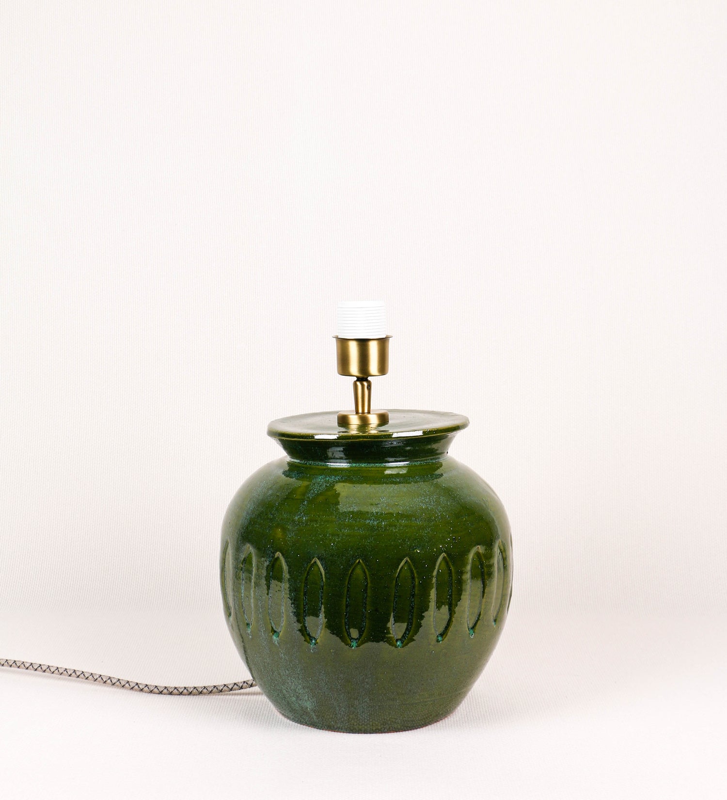 Candeeiro de mesa com um design rústico e sofisticado. A base do candeeiro é de barro vidrado de tom verde vidro. A forma da base é arredondada com uma borda superior mais estreita, lembrando um vaso tradicional. A superfície da base é decorada com um padrão de relevos ovais. O suporte da lâmpada é em latão escovado e é manipulável. O cabo é revestido têxtil com um padrão trançado de linhas castanhas e beges, com interruptor e ficha transparentes.