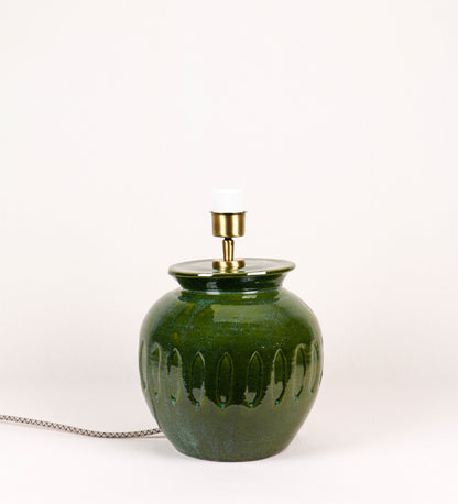 Candeeiro de mesa com um design rústico e sofisticado. A base do candeeiro é de barro vidrado de tom verde vidro. A forma da base é arredondada com uma borda superior mais estreita, lembrando um vaso tradicional. A superfície da base é decorada com um padrão de relevos ovais. O suporte da lâmpada é em latão escovado e é manipulável. O cabo é revestido têxtil com um padrão trançado de linhas castanhas e beges, com interruptor e ficha transparentes.
