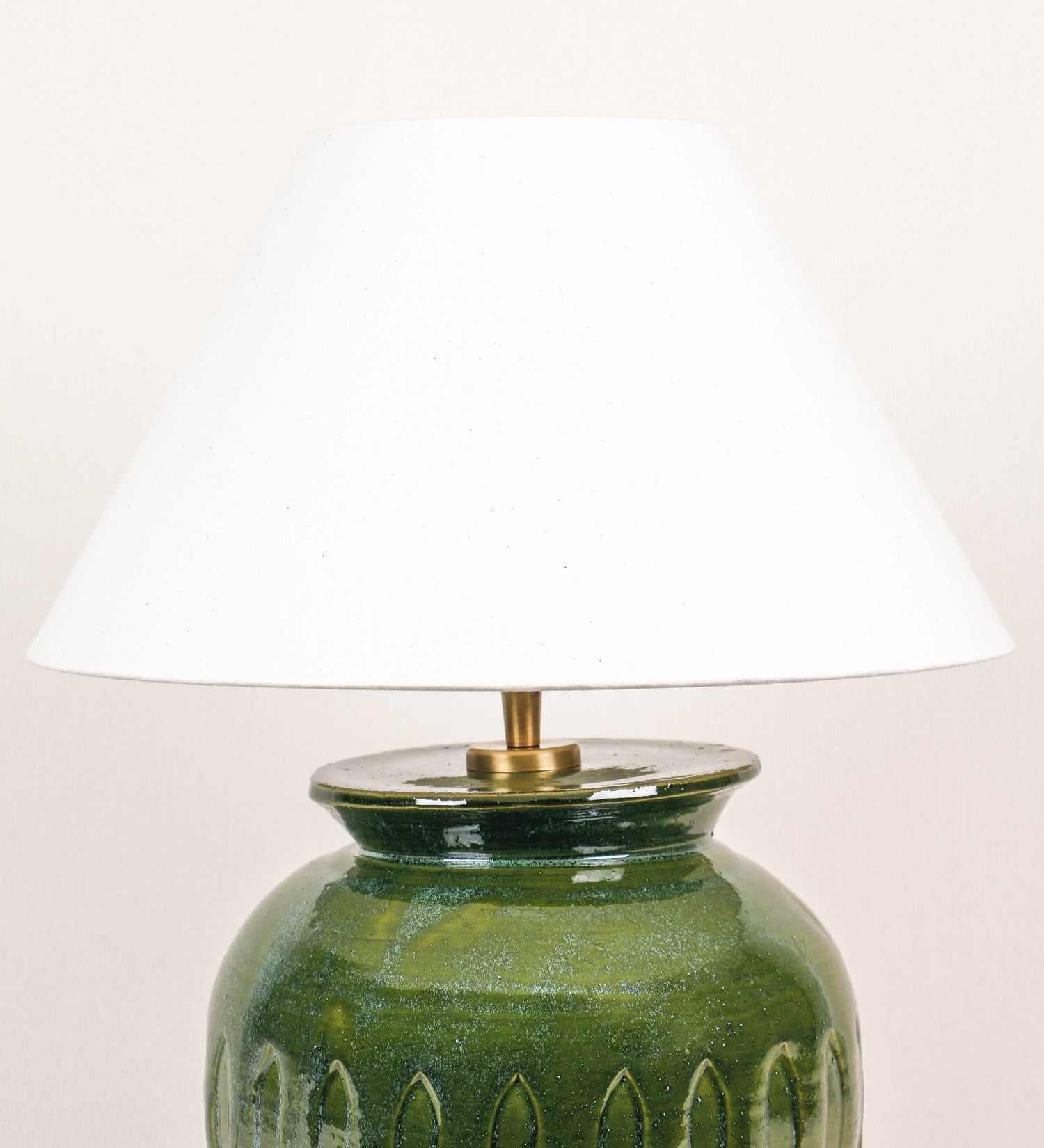 Candeeiro de mesa com um design rústico e sofisticado. A base do candeeiro é de barro vidrado de tom verde vidro. A forma da base é arredondada com uma borda superior mais estreita, lembrando um vaso tradicional. A superfície da base é decorada com um padrão de relevos ovais. O suporte da lâmpada é de latão escovado e é manipulável. O cabo é revestido têxtil com um padrão trançado de linhas castanhas e beges, com interruptor e ficha transparentes. O abatjour é cónico, em algodão, de tom bege claro.
