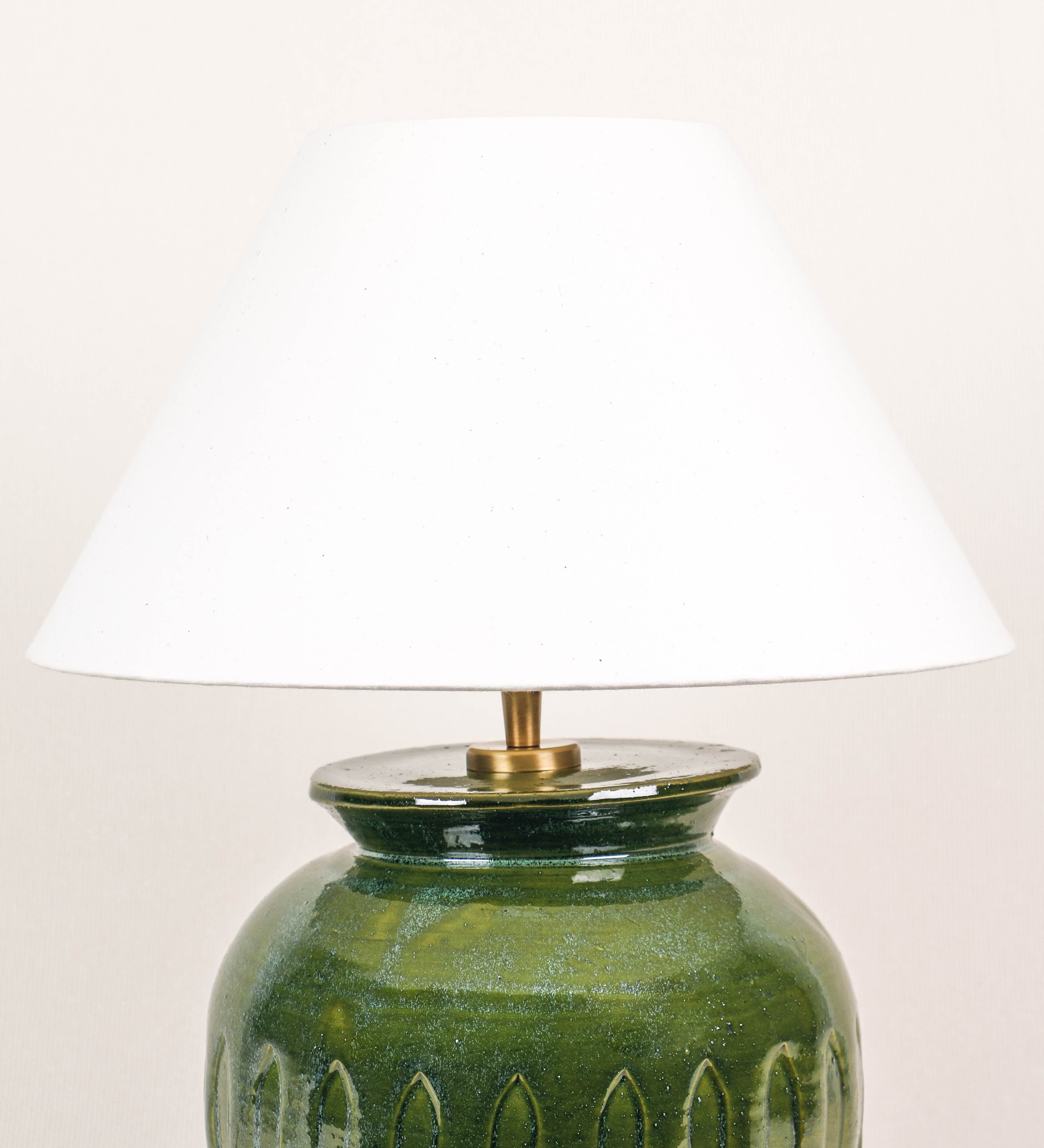 Candeeiro de mesa com um design rústico e sofisticado. A base do candeeiro é de barro vidrado de tom verde vidro. A forma da base é arredondada com uma borda superior mais estreita, lembrando um vaso tradicional. A superfície da base é decorada com um padrão de relevos ovais. O suporte da lâmpada é de latão escovado e é manipulável. O cabo é revestido têxtil com um padrão trançado de linhas castanhas e beges, com interruptor e ficha transparentes. O abatjour é cónico, em algodão, de tom bege claro.