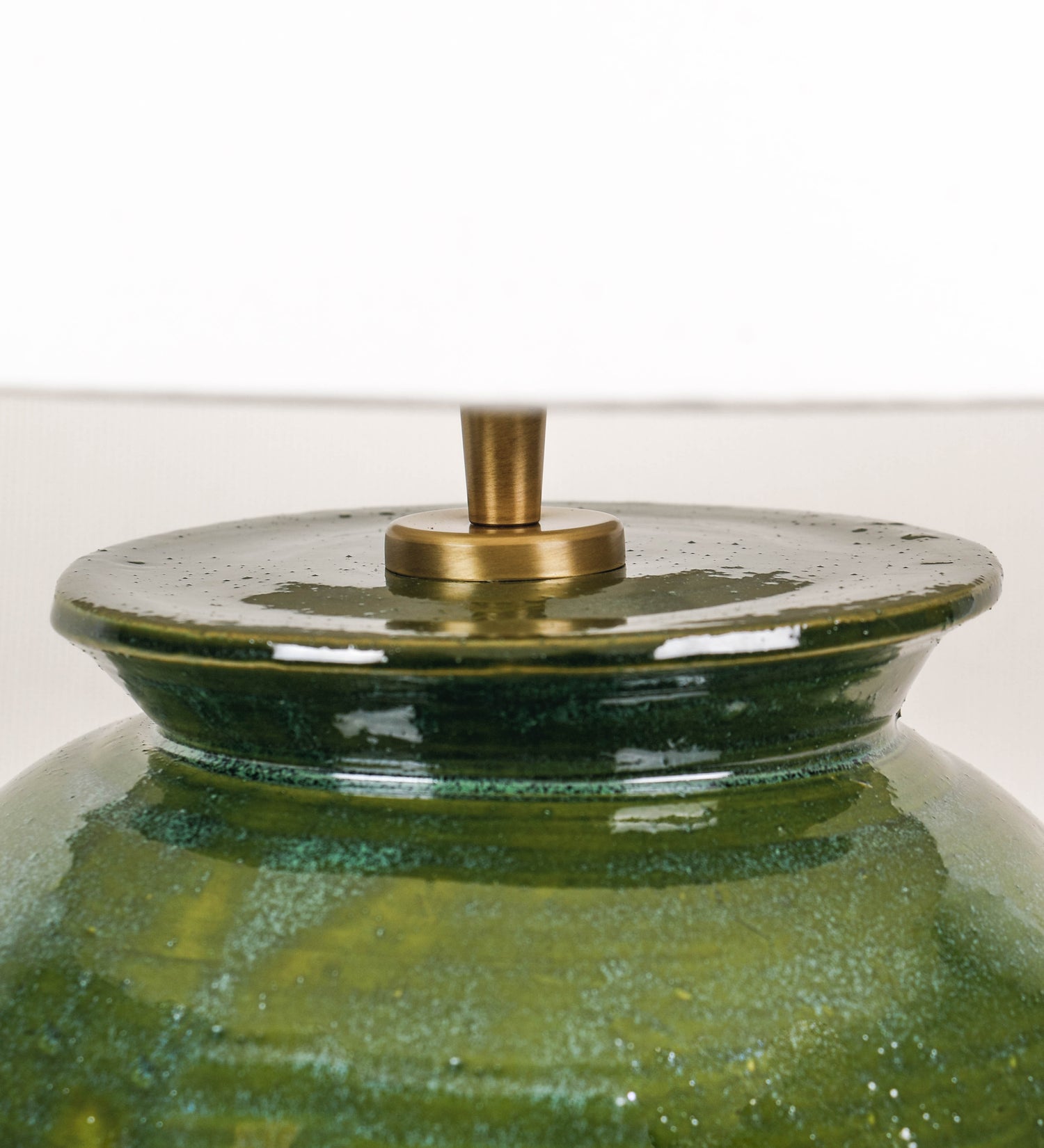 Candeeiro de mesa com um design rústico e sofisticado. A base do candeeiro é de barro vidrado de tom verde vidro. A forma da base é arredondada com uma borda superior mais estreita, lembrando um vaso tradicional. A superfície da base é decorada com um padrão de relevos ovais. O suporte da lâmpada é de latão escovado e é manipulável. O cabo é revestido têxtil com um padrão trançado de linhas castanhas e beges, com interruptor e ficha transparentes. O abatjour é cónico, em algodão, de tom bege claro.