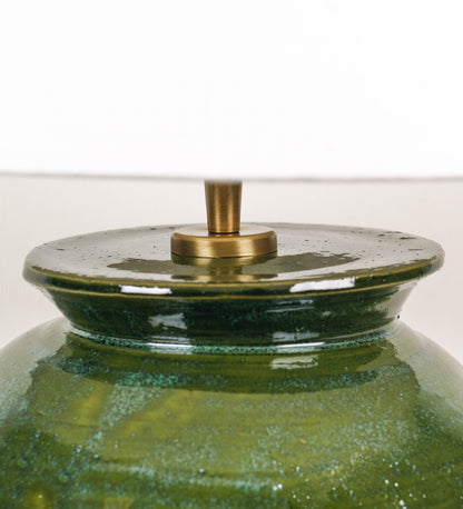 Candeeiro de mesa com um design rústico e sofisticado. A base do candeeiro é de barro vidrado de tom verde vidro. A forma da base é arredondada com uma borda superior mais estreita, lembrando um vaso tradicional. A superfície da base é decorada com um padrão de relevos ovais. O suporte da lâmpada é de latão escovado e é manipulável. O cabo é revestido têxtil com um padrão trançado de linhas castanhas e beges, com interruptor e ficha transparentes. O abatjour é cónico, em algodão, de tom bege claro.