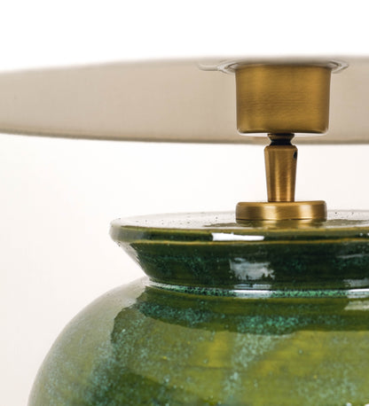 Candeeiro de mesa com um design rústico e sofisticado. A base do candeeiro é de barro vidrado de tom verde vidro. A forma da base é arredondada com uma borda superior mais estreita, lembrando um vaso tradicional. A superfície da base é decorada com um padrão de relevos ovais. O suporte da lâmpada é de latão escovado e é manipulável. O cabo é revestido têxtil com um padrão trançado de linhas castanhas e beges, com interruptor e ficha transparentes. O abatjour é cónico, em algodão, de tom bege claro.