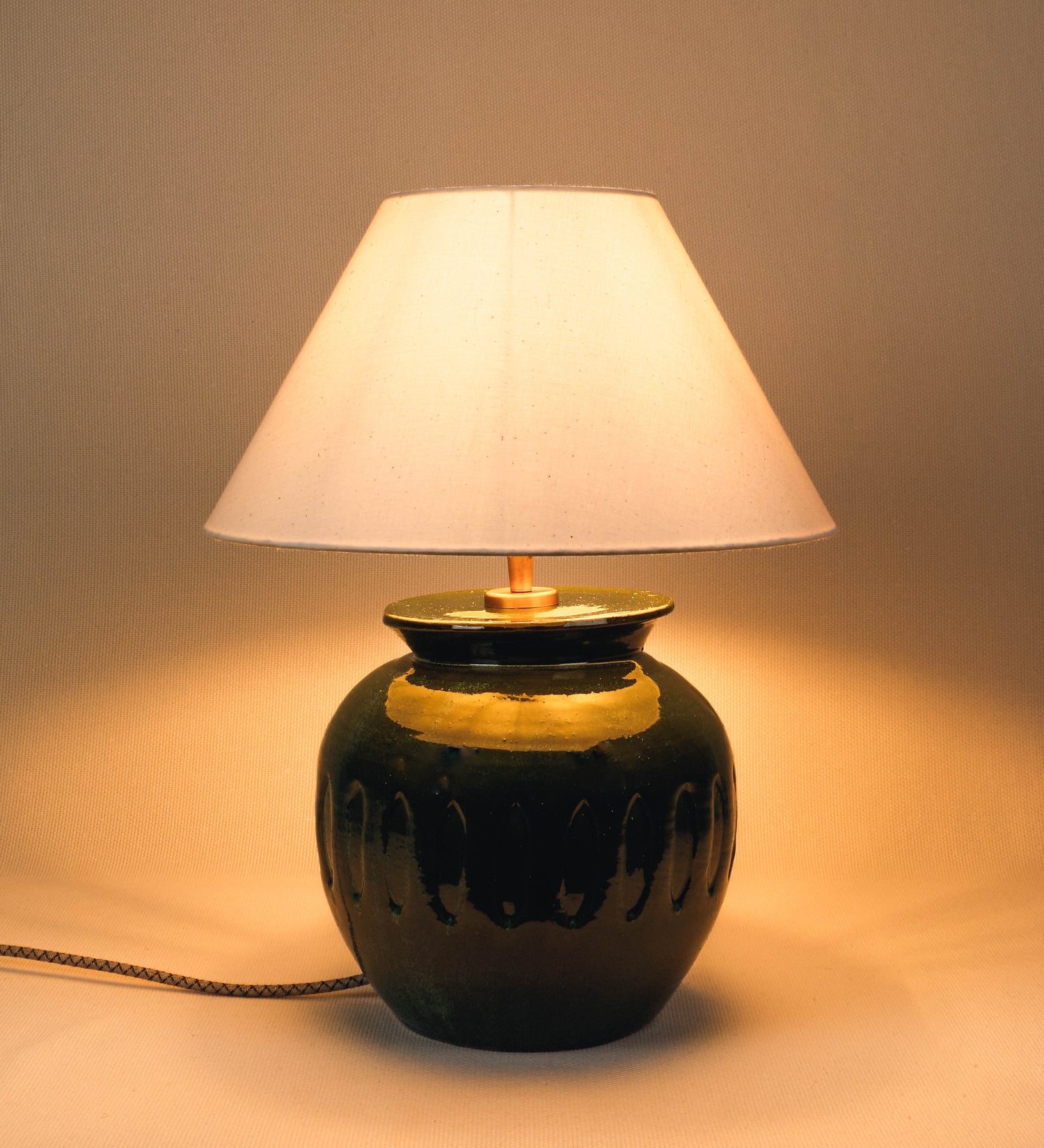 Candeeiro de mesa com um design rústico e sofisticado. A base do candeeiro é de barro vidrado de tom verde vidro. A forma da base é arredondada com uma borda superior mais estreita, lembrando um vaso tradicional. A superfície da base é decorada com um padrão de relevos ovais. O suporte da lâmpada é de latão escovado e é manipulável. O cabo é revestido têxtil com um padrão trançado de linhas castanhas e beges, com interruptor e ficha transparentes. O abatjour é cónico, em algodão, de tom bege claro.